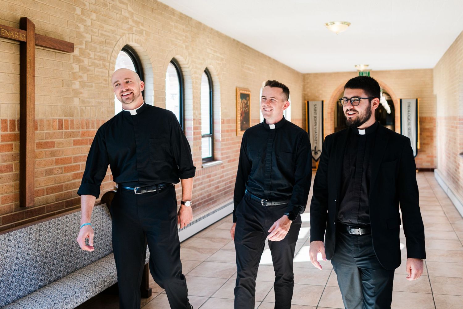 Three Catholic priests in black clerical attire walk and smile together along a sunlit church corridor.