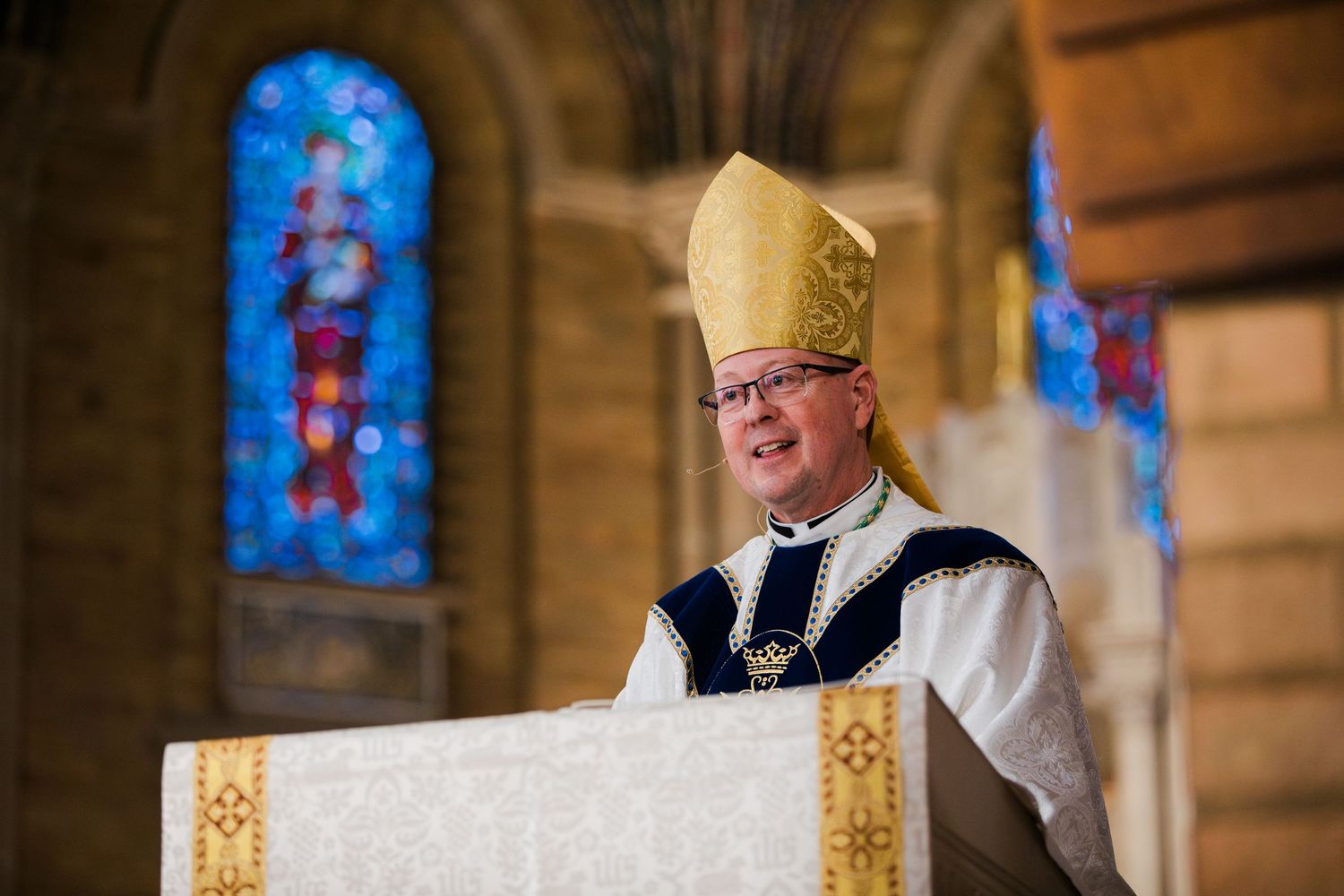 Bishop in golden mitre delivers sermon at ornate church pulpit with colorful stained glass windows behind him.