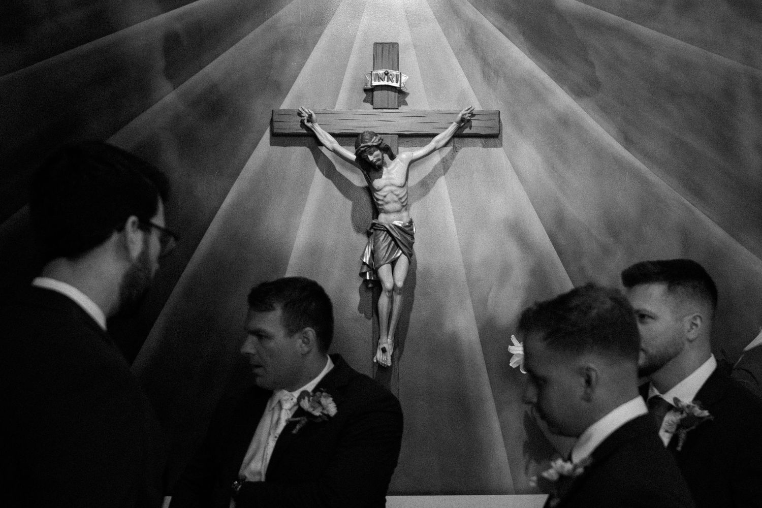 Black and white photo of groomsmen standing beneath a crucifix in a church during a wedding ceremony.