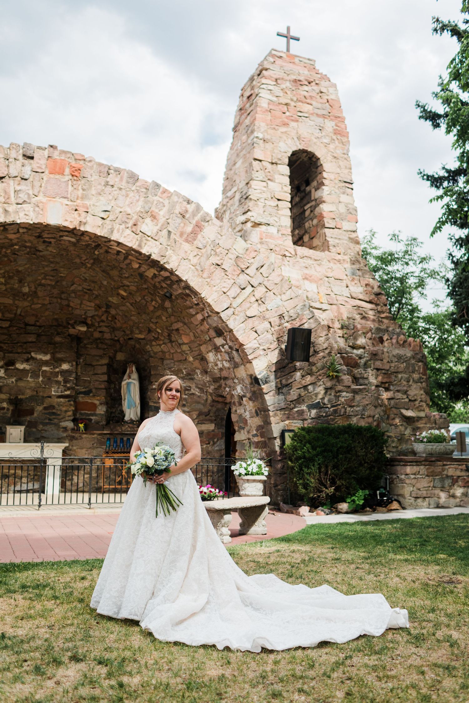 Bride in lace gown poses before a rustic stone grotto with cross and Mary statue outdoors.