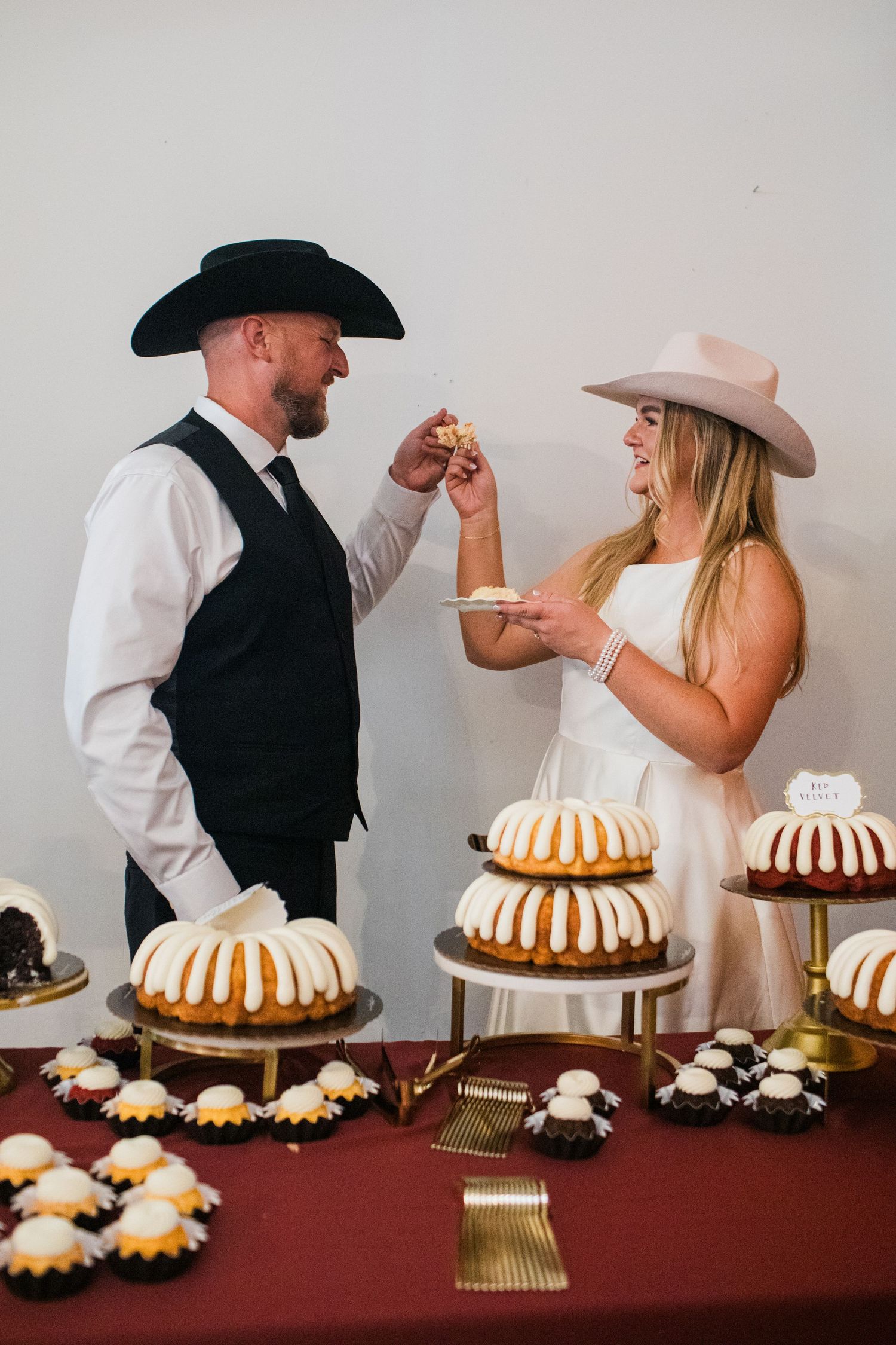 Bride and groom in cowboy hats cutting wedding cake at a dessert table filled with bundt cakes.