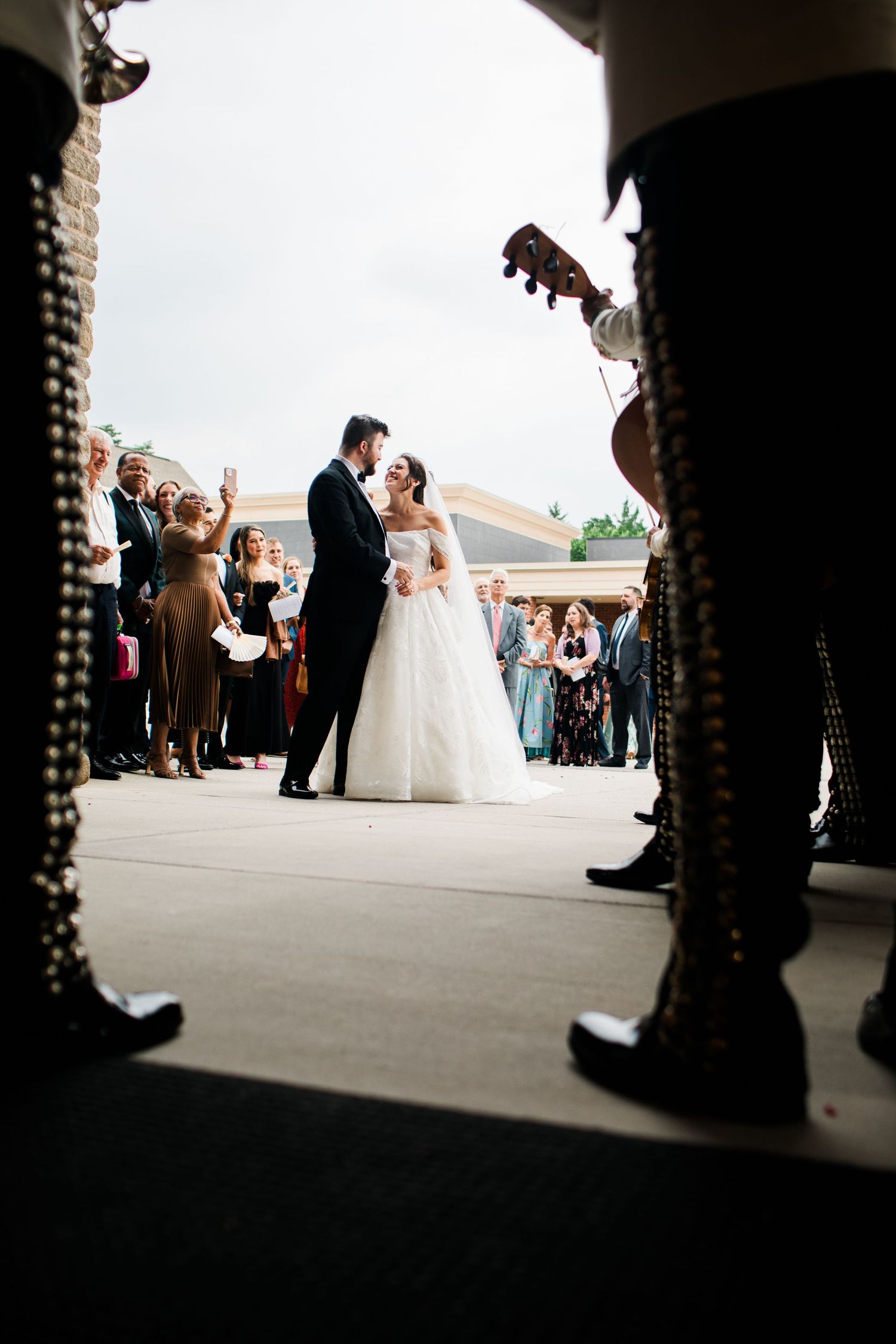 Bride and groom share first dance outdoors while mariachi band performs for wedding guests gathered around them.