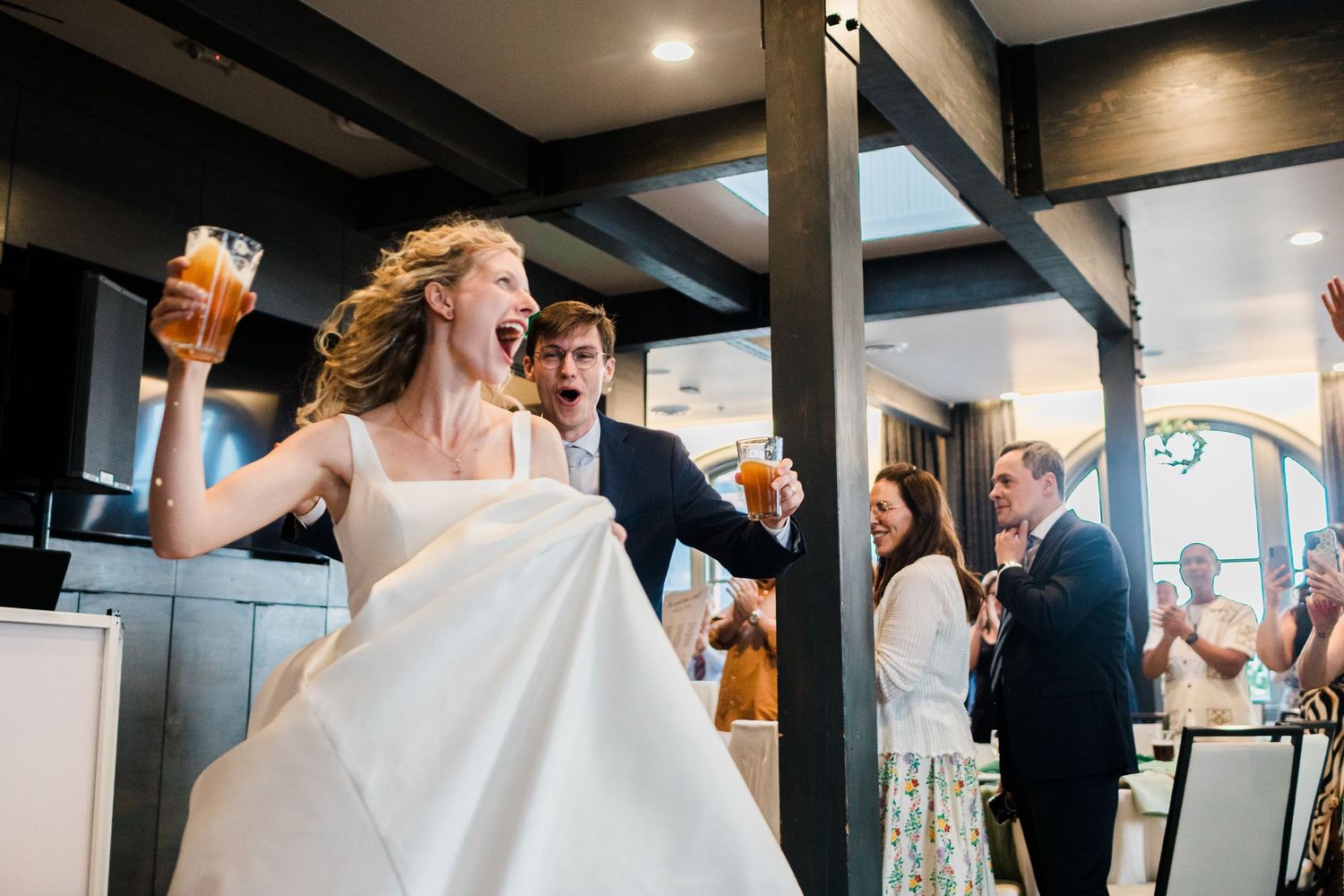 Joyful bride in white gown raises beer glass with groom during lively wedding reception entrance.