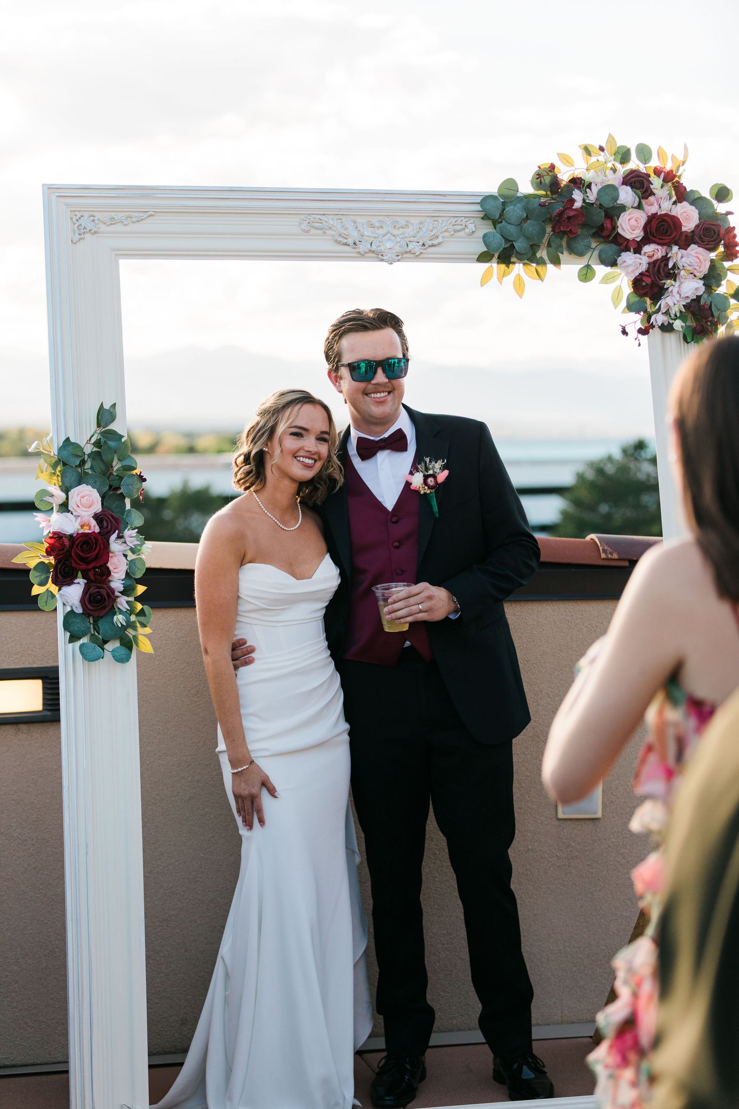 Bride and groom pose in decorative white frame with floral accents at outdoor rooftop wedding reception.