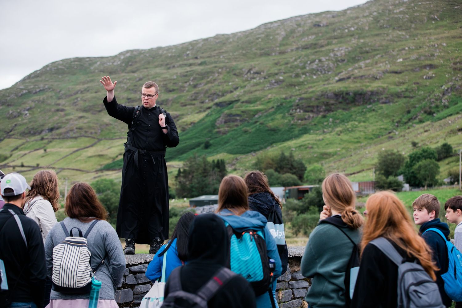 Priest in black robes addresses a group of students with green hills in the background outdoors.