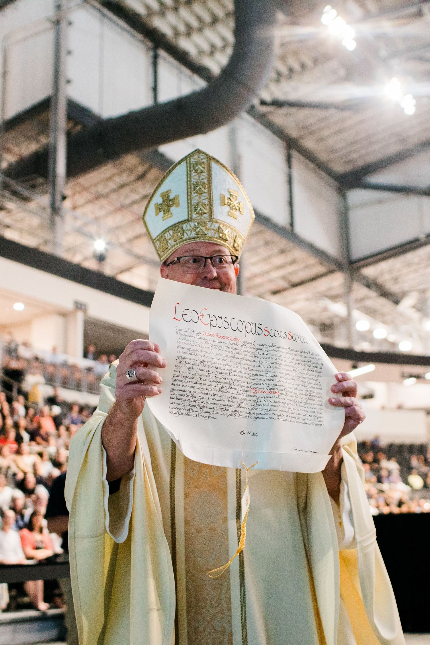 Catholic bishop in gold vestments and mitre holding up official papal document during ordination Mass.