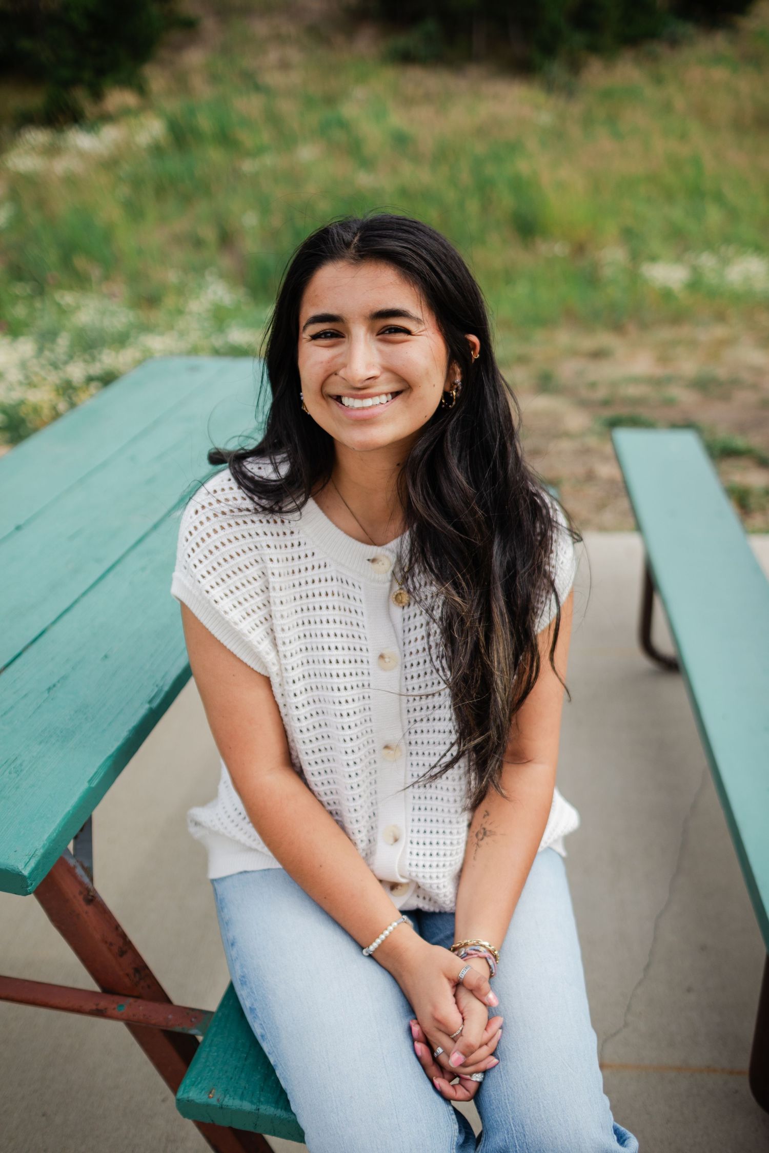 A person in a white crochet top and light jeans sits casually on a green picnic bench outdoors.