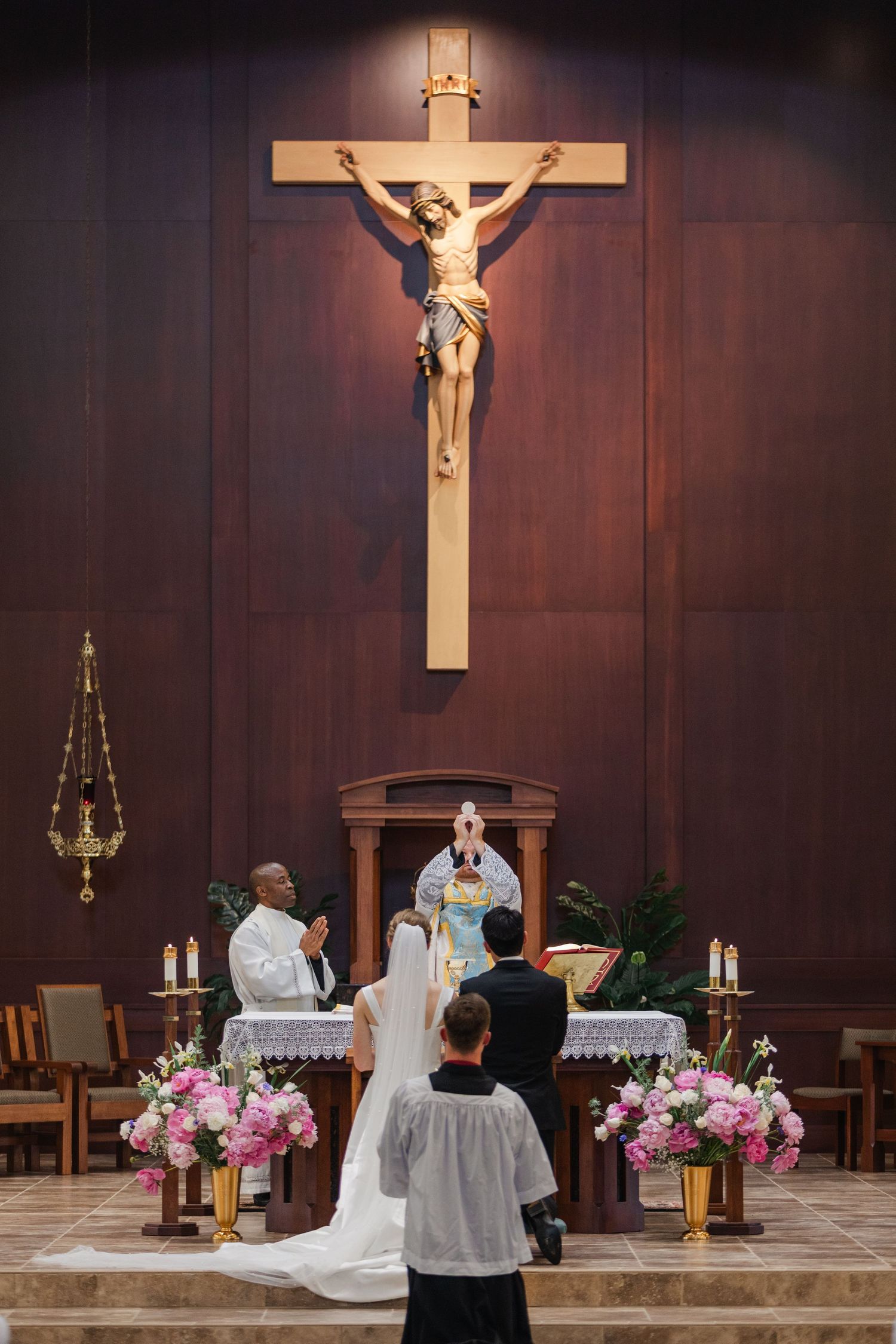 A Catholic mass ceremony at the altar with clergy members performing religious rituals surrounded by floral arrangements.