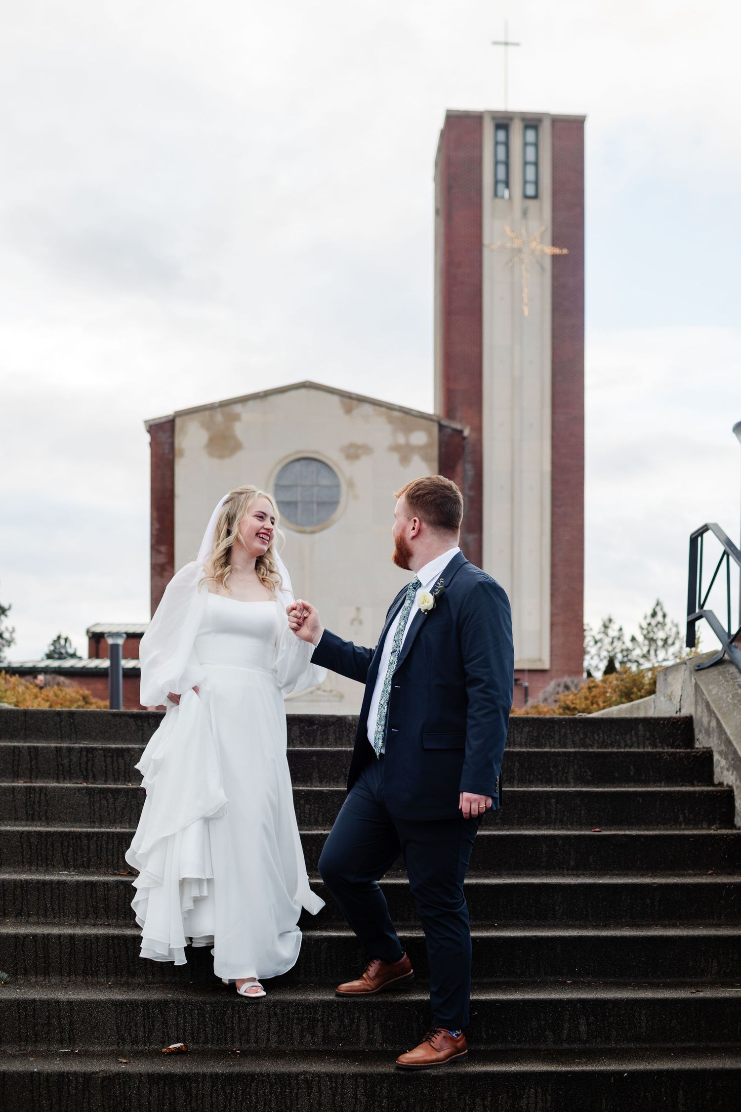 A bride and groom share a candid moment on concrete steps outside a building with tall towers.