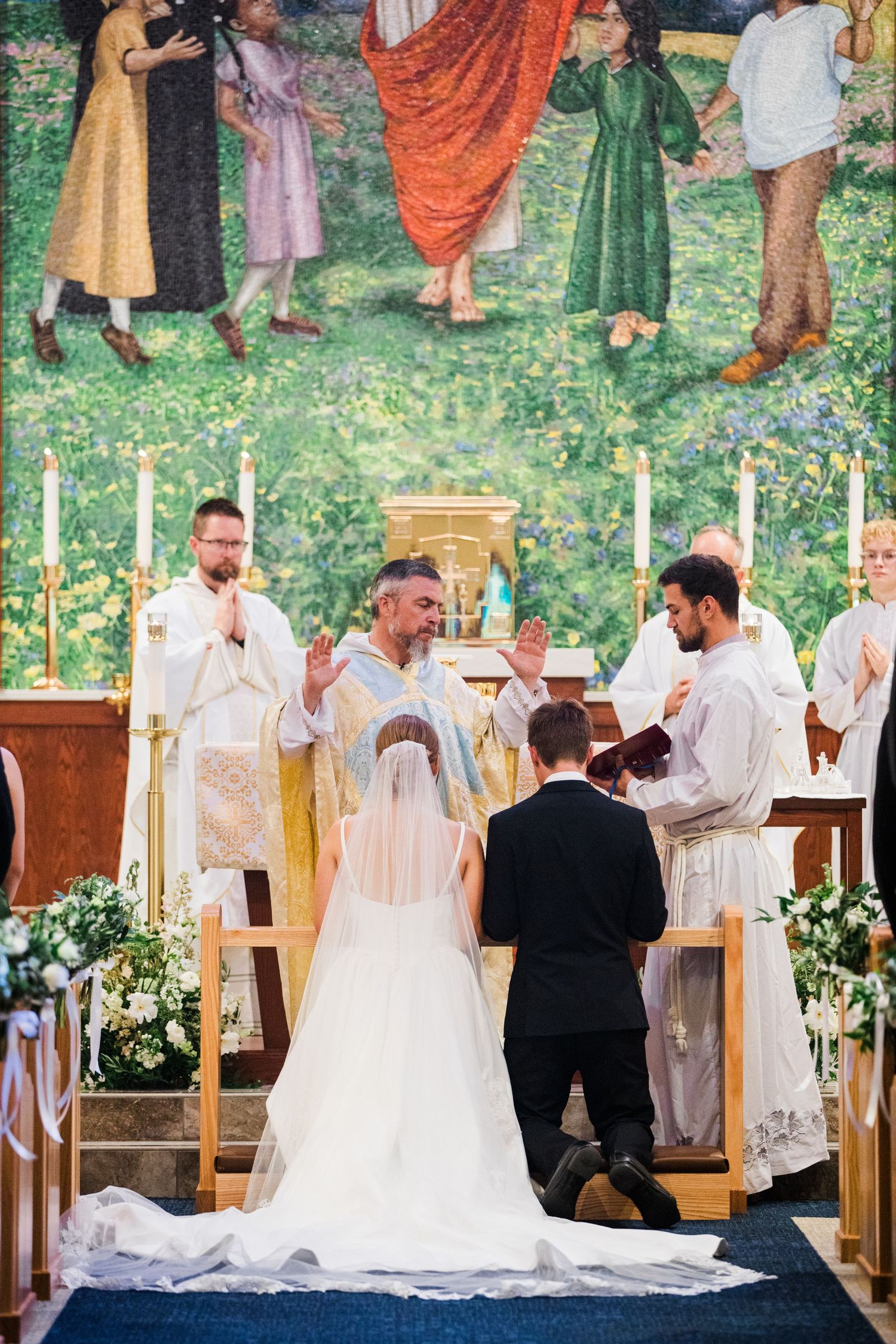 Wedding ceremony in progress at a church with decorative wall murals and floral arrangements.