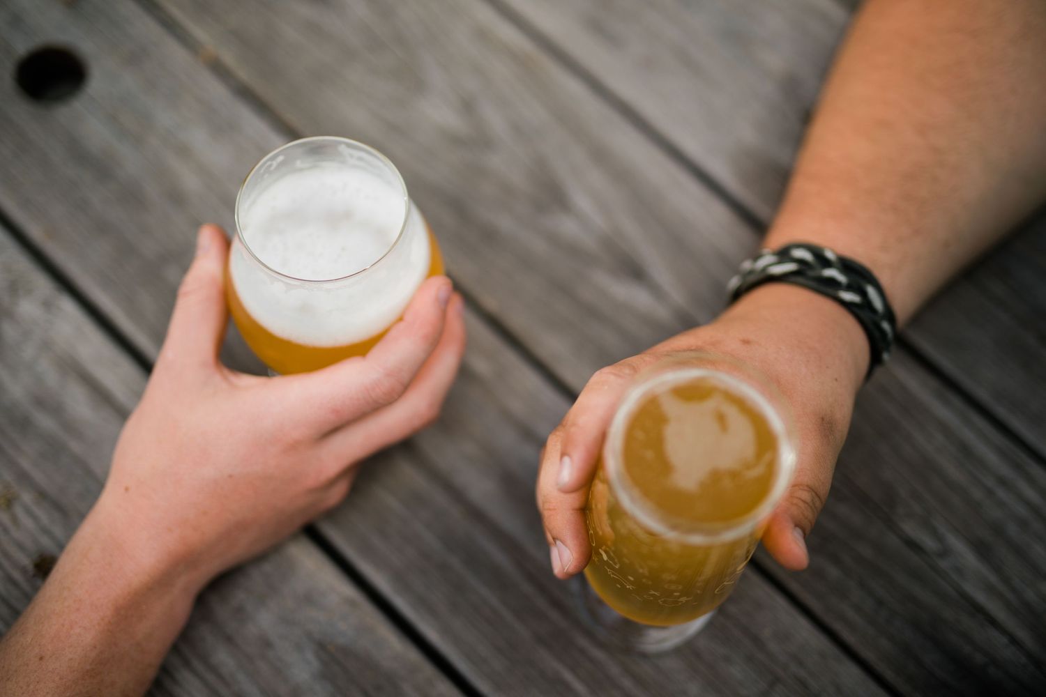 Multiple hands holding glasses of golden beer over weathered wooden deck planks.