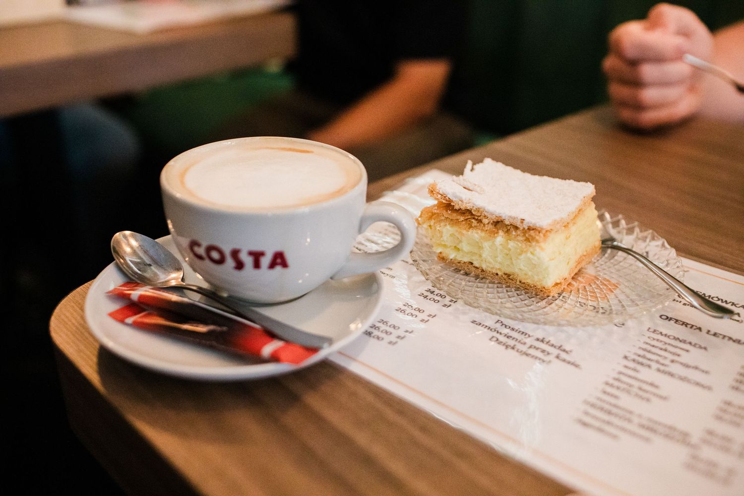 Costa Coffee cappuccino with frothy milk beside a slice of vanilla cream cake on a wooden cafe table.