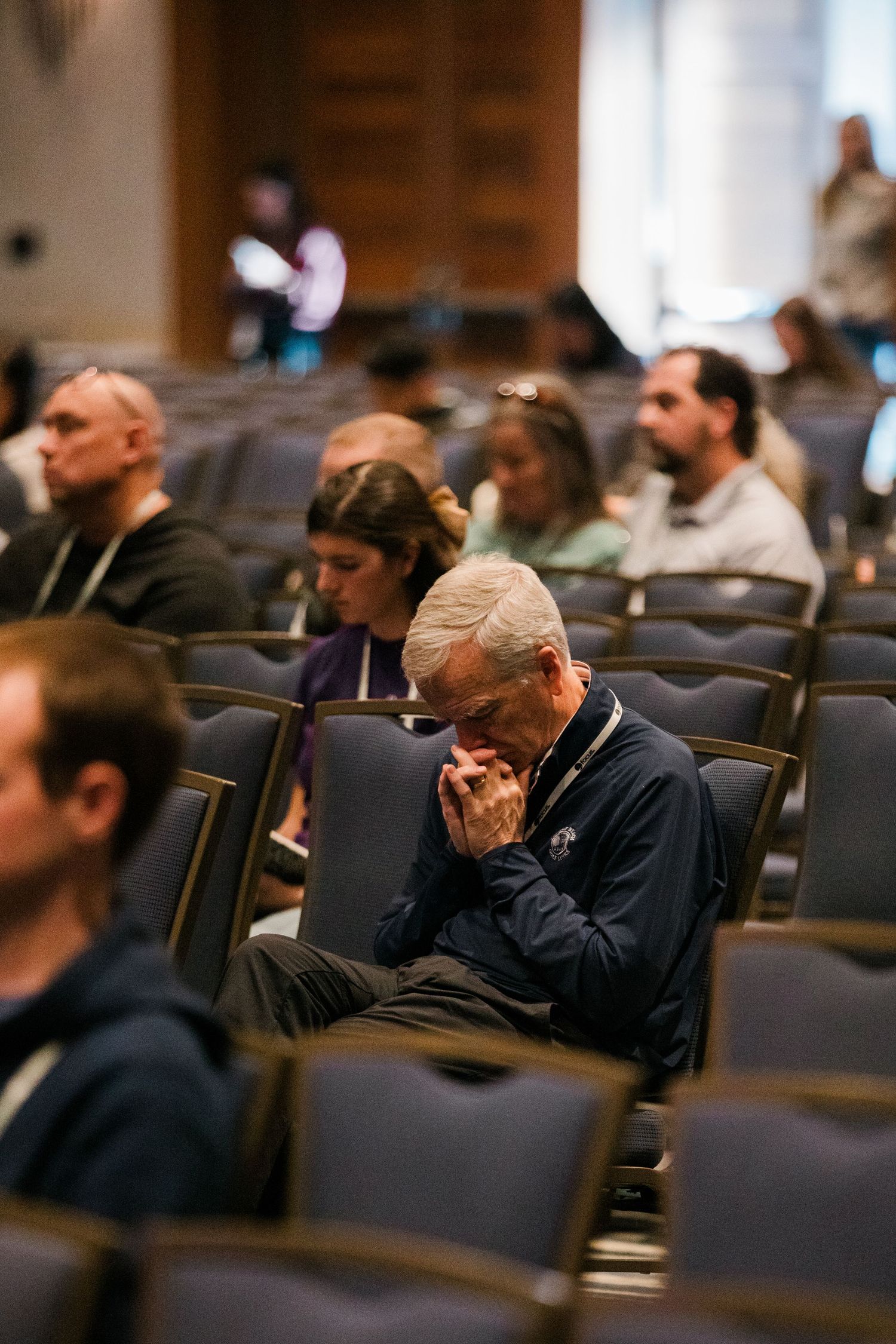 People sit in rows of chairs at a conference or meeting with one attendee bowing their head in contemplation.