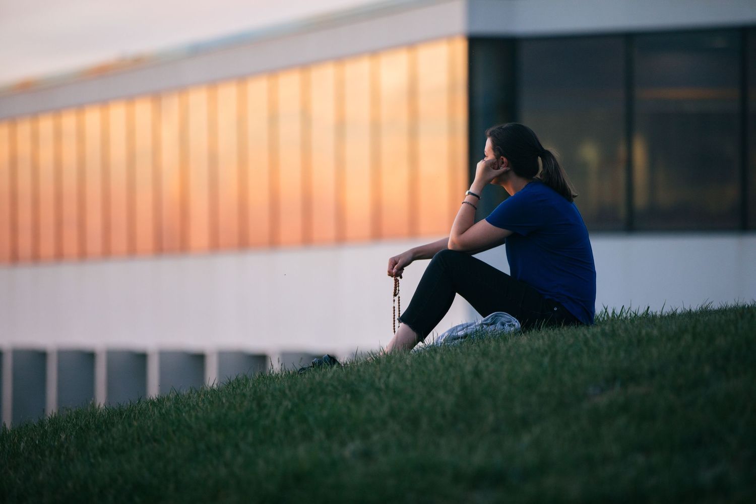A person sits alone on a grassy slope against a building at sunset creating a contemplative silhouette.