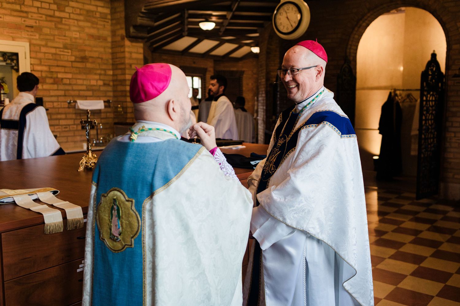 Two Catholic bishops in ornate vestments and miters converse warmly inside a church during a religious ceremony.