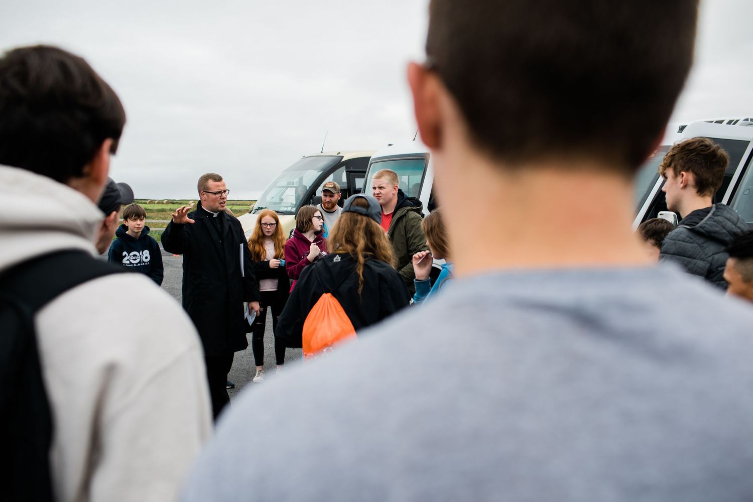 Group of young people gathered outdoors near a bus listening to a speaker on an overcast day.