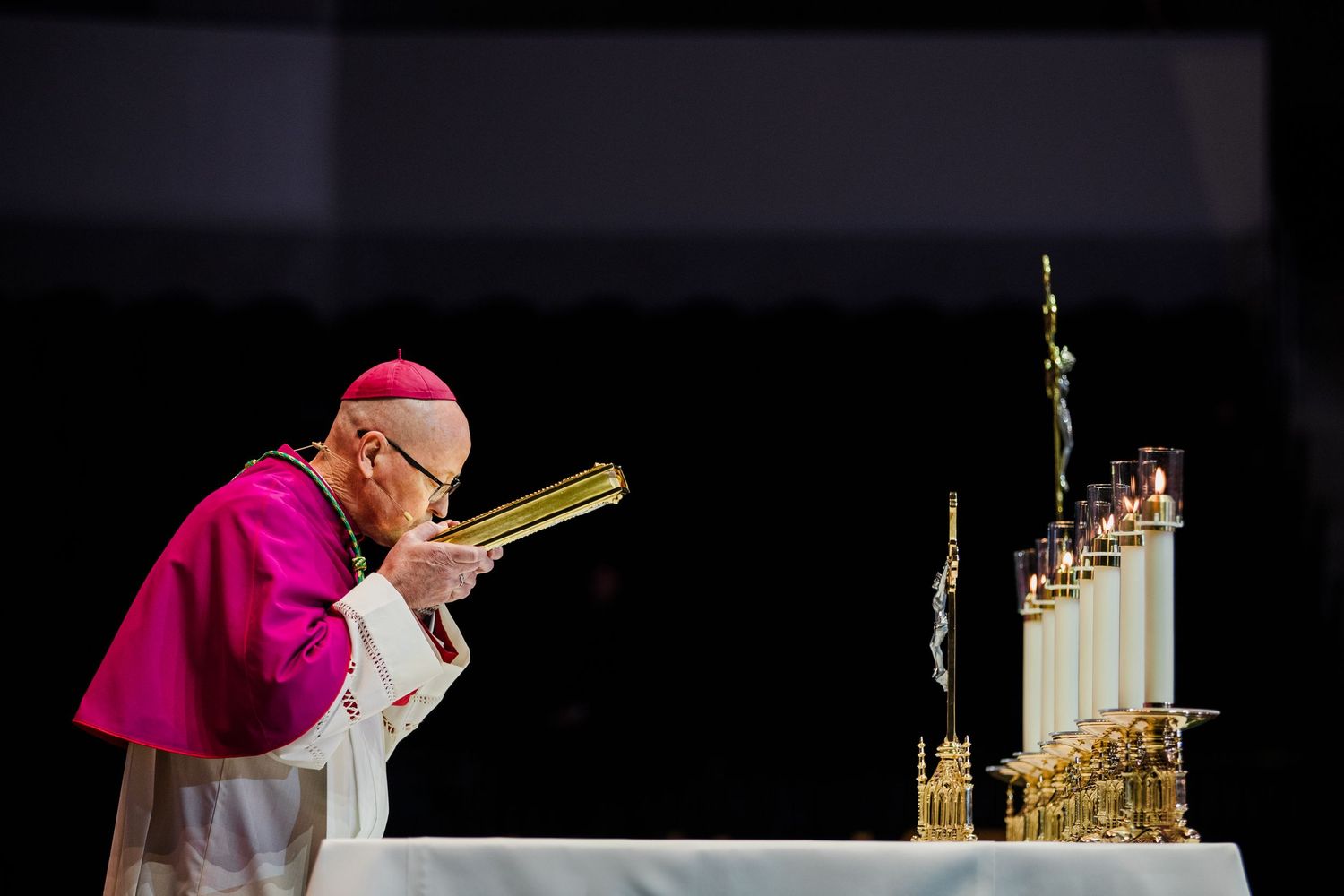 Bishop in pink vestments bows reverently at candlelit altar during solemn Catholic Mass ceremony.