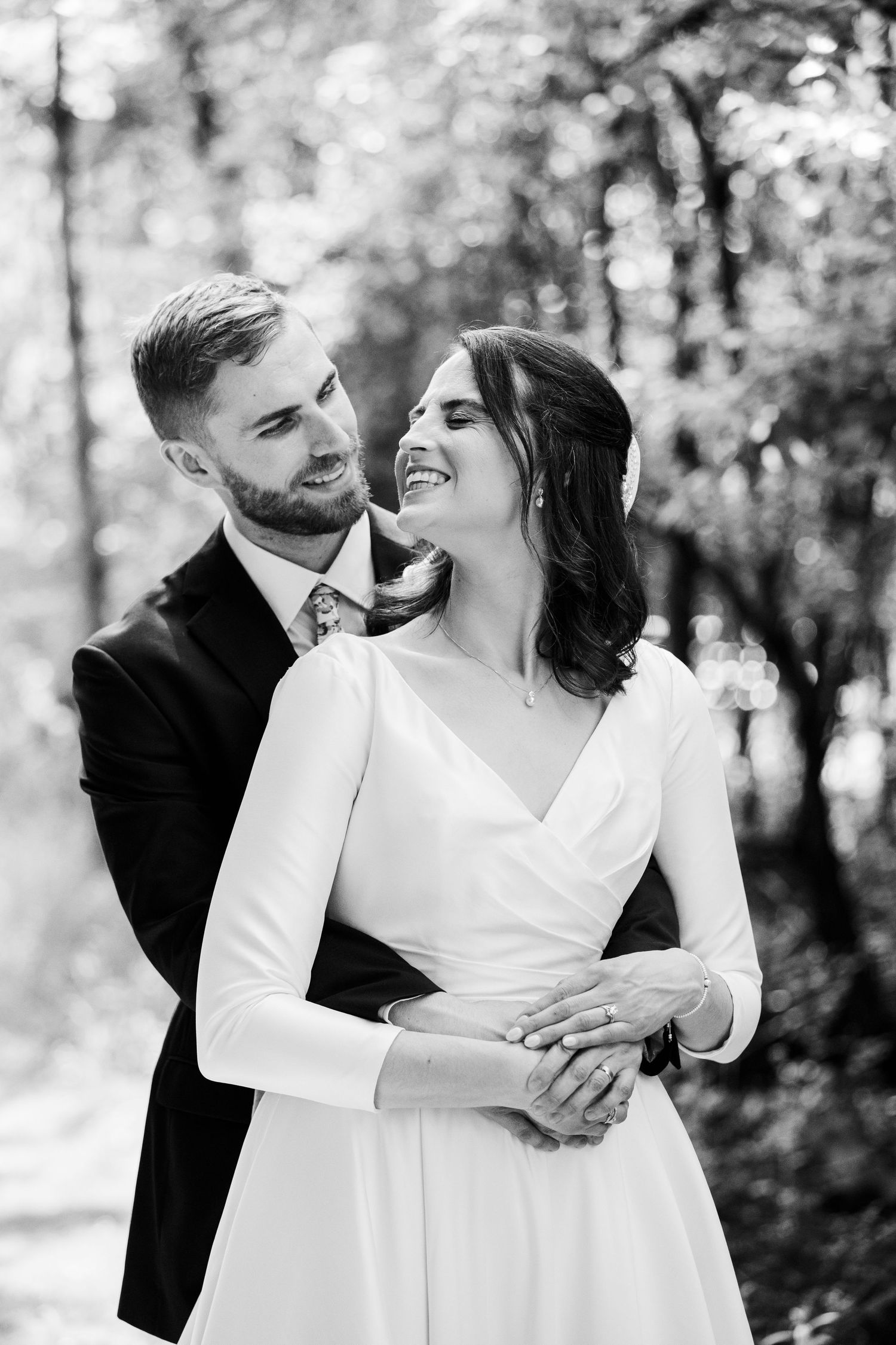 Black and white portrait of groom embracing laughing bride in white dress outdoors among trees.