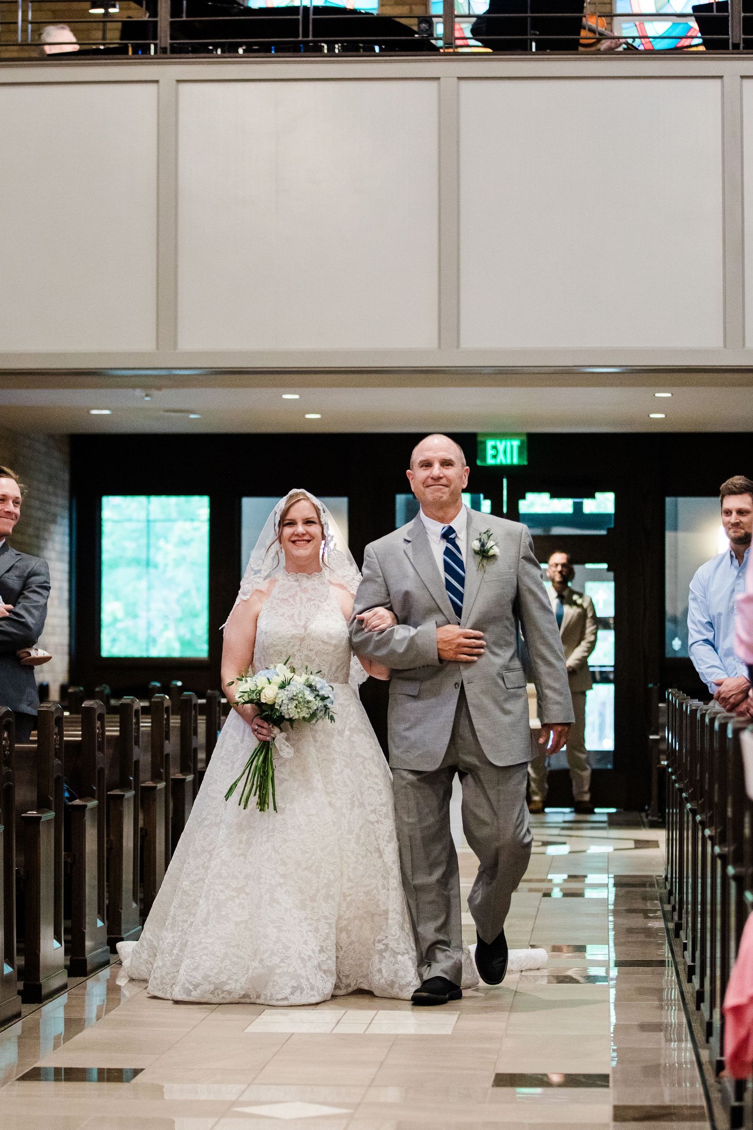 Bride in lace gown walks down church aisle with escort, holding white bouquet, guests watching from wooden pews.