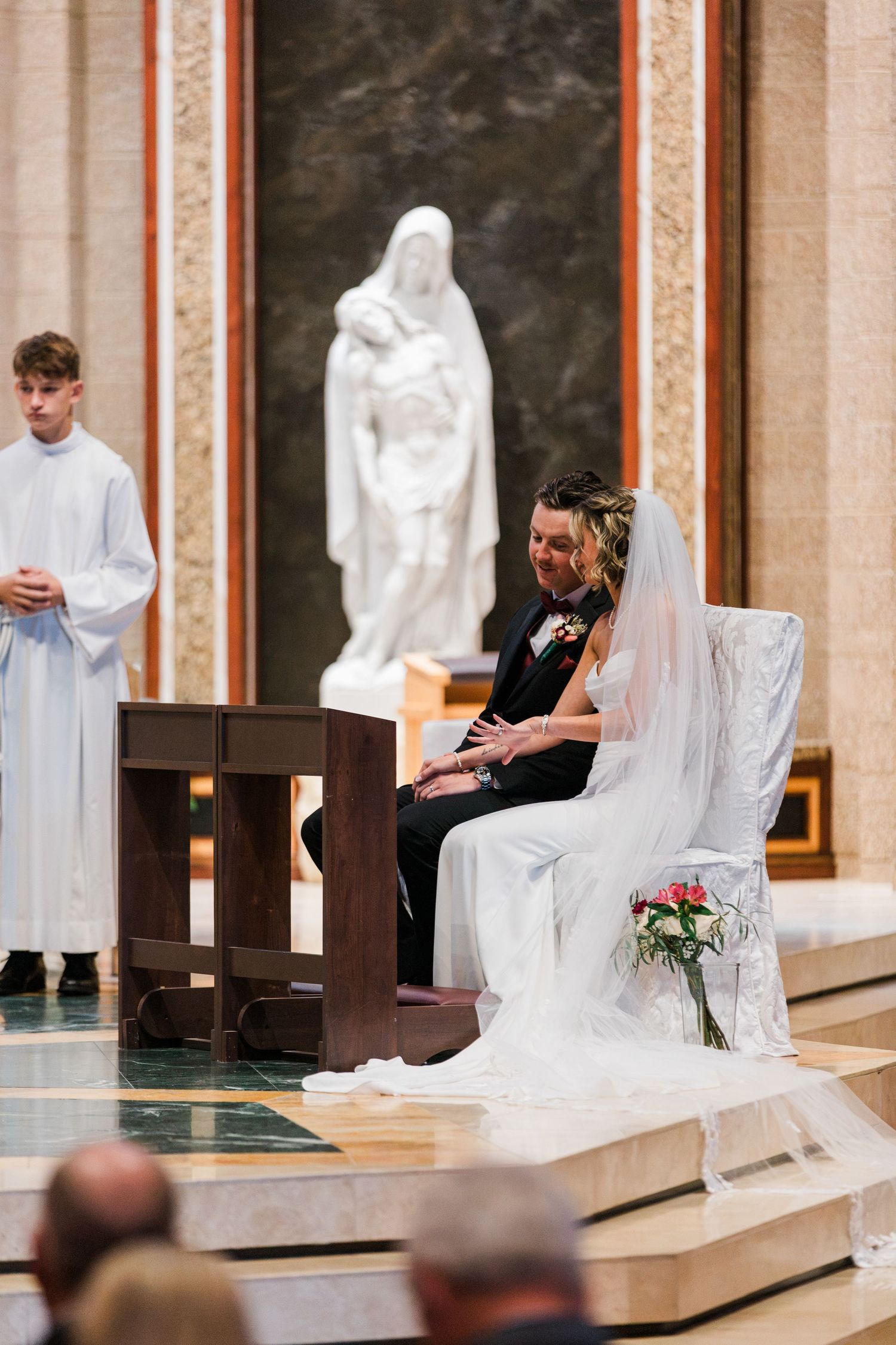 Bride and groom sit together at altar during Catholic wedding ceremony with white statue behind.