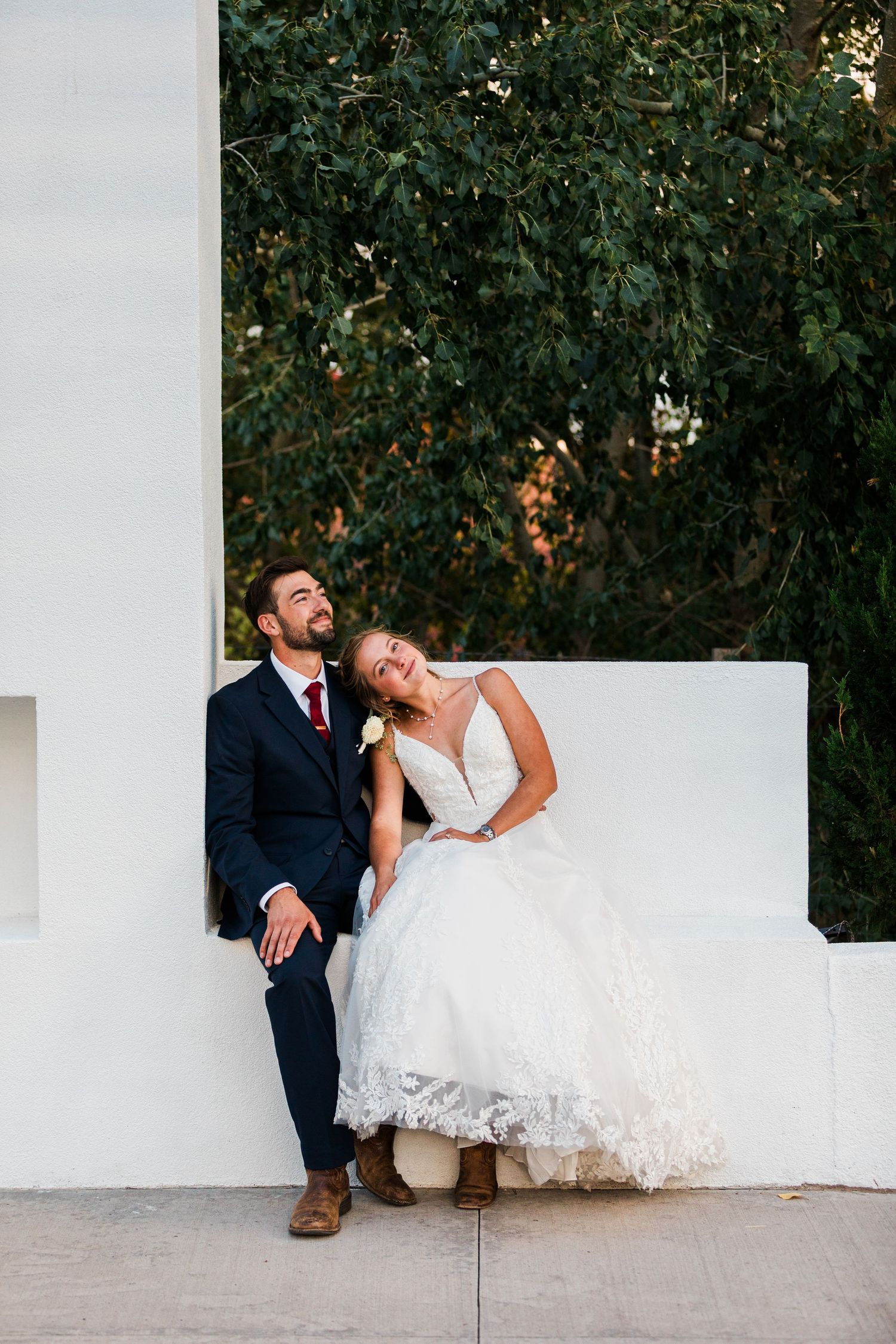 A joyful bride in lace gown leans on groom in navy suit, sitting together outdoors against a white wall.