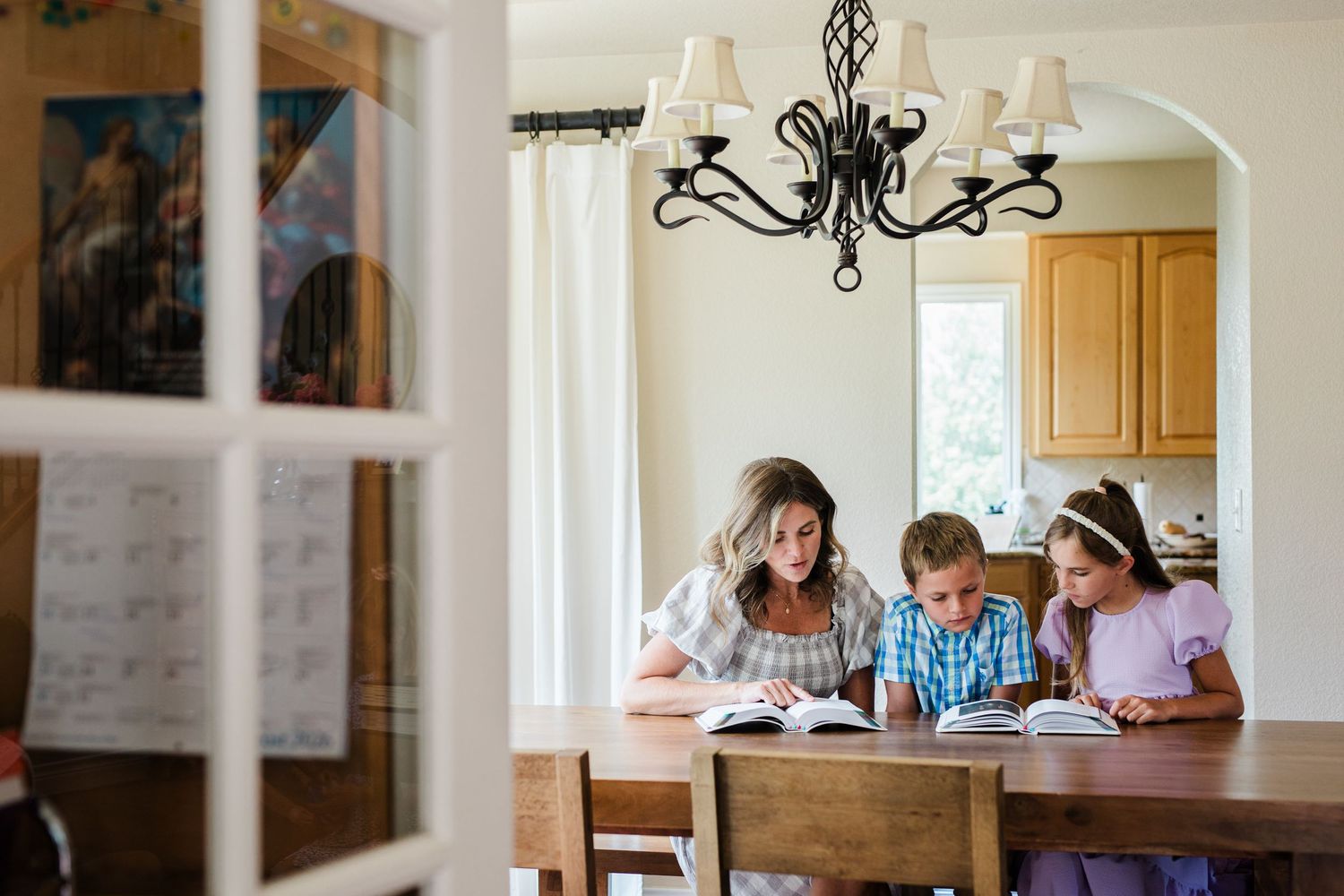 A mother homeschooling her two children at a wooden dining table, reviewing books together in a warm home setting.