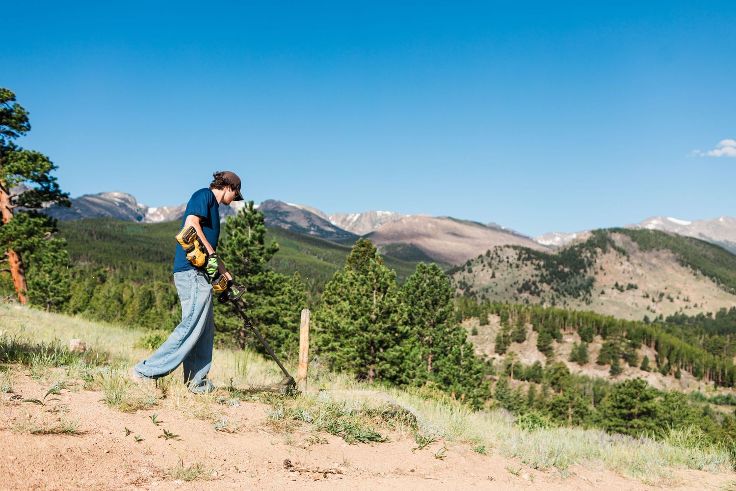 Person using a metal detector on a mountain trail with scenic Rocky Mountain peaks and pine trees in background.