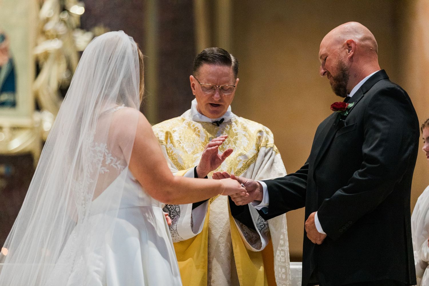 Bride and groom exchange rings during Catholic wedding ceremony with priest in golden vestments.