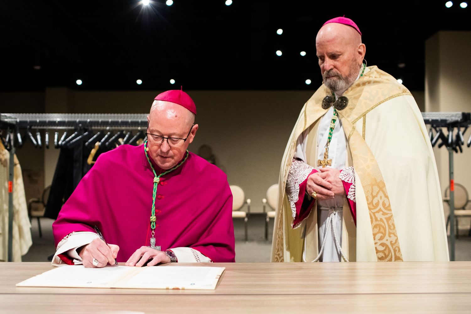 Catholic bishop in pink vestments signing official documents at table during formal church ceremony.