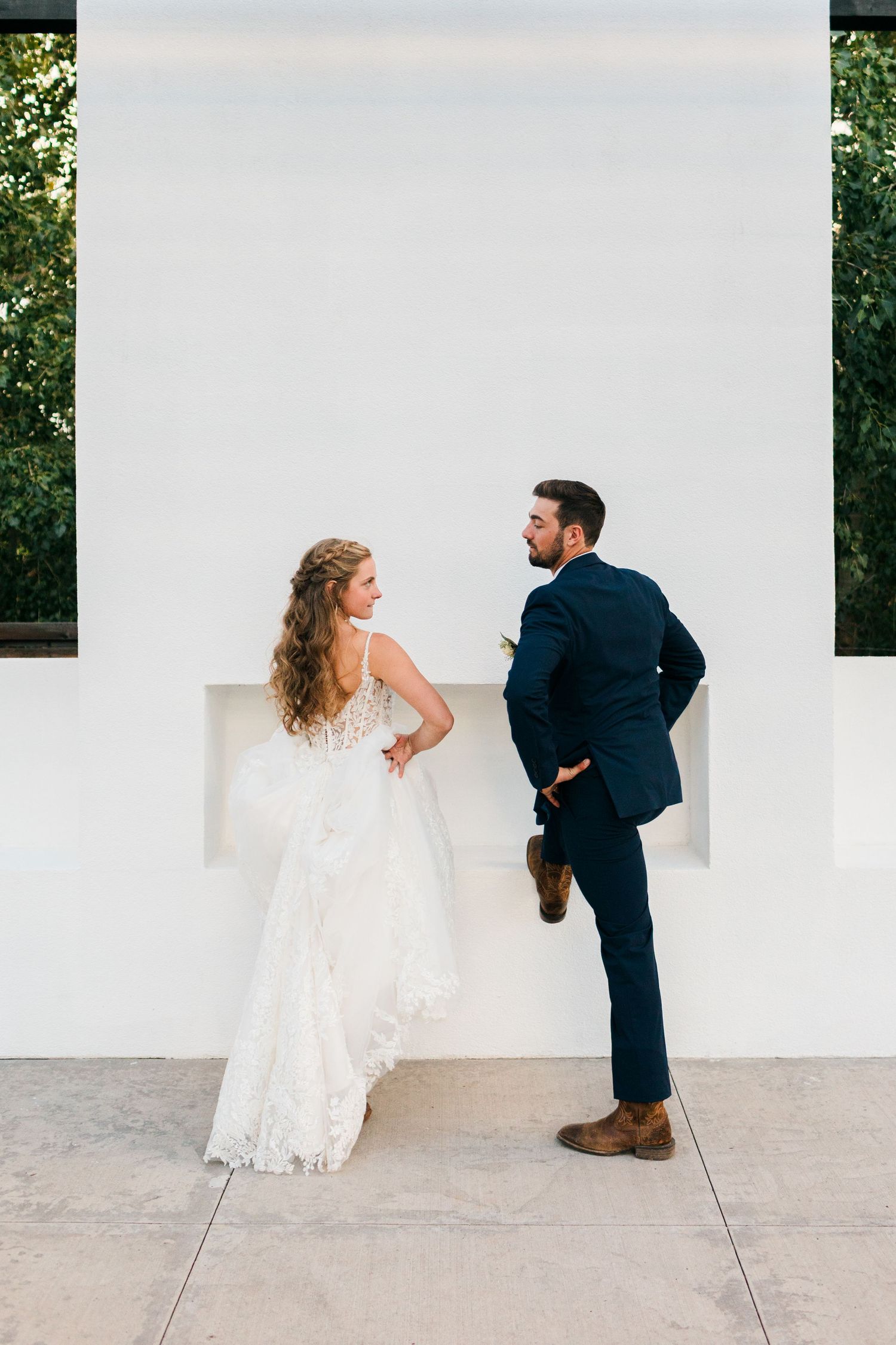 Bride in lace gown and groom in navy suit share a playful moment against a white wall outdoors.