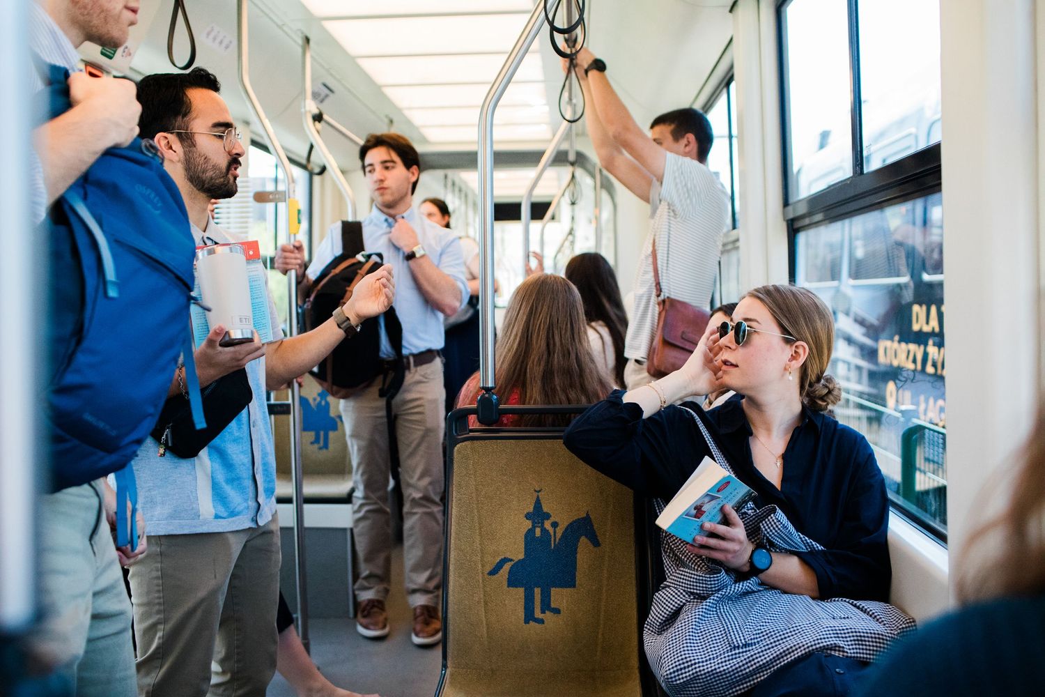 Passengers socializing on a busy tram, with a woman reading a book and a blue dragon logo on a seat.