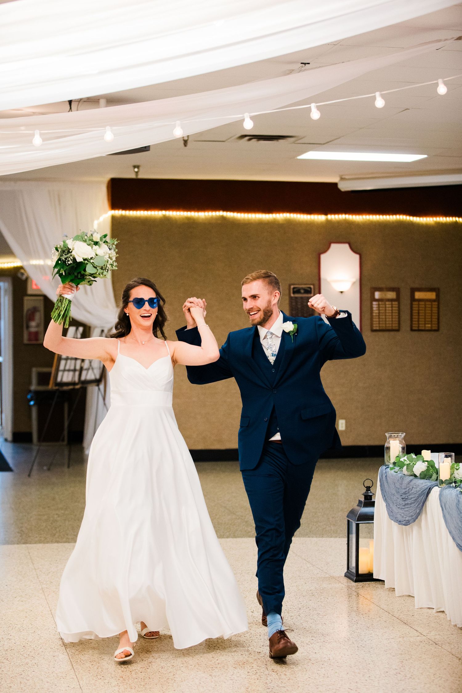 Joyful newlyweds wearing sunglasses make grand entrance at wedding reception hall, bride holding bouquet.