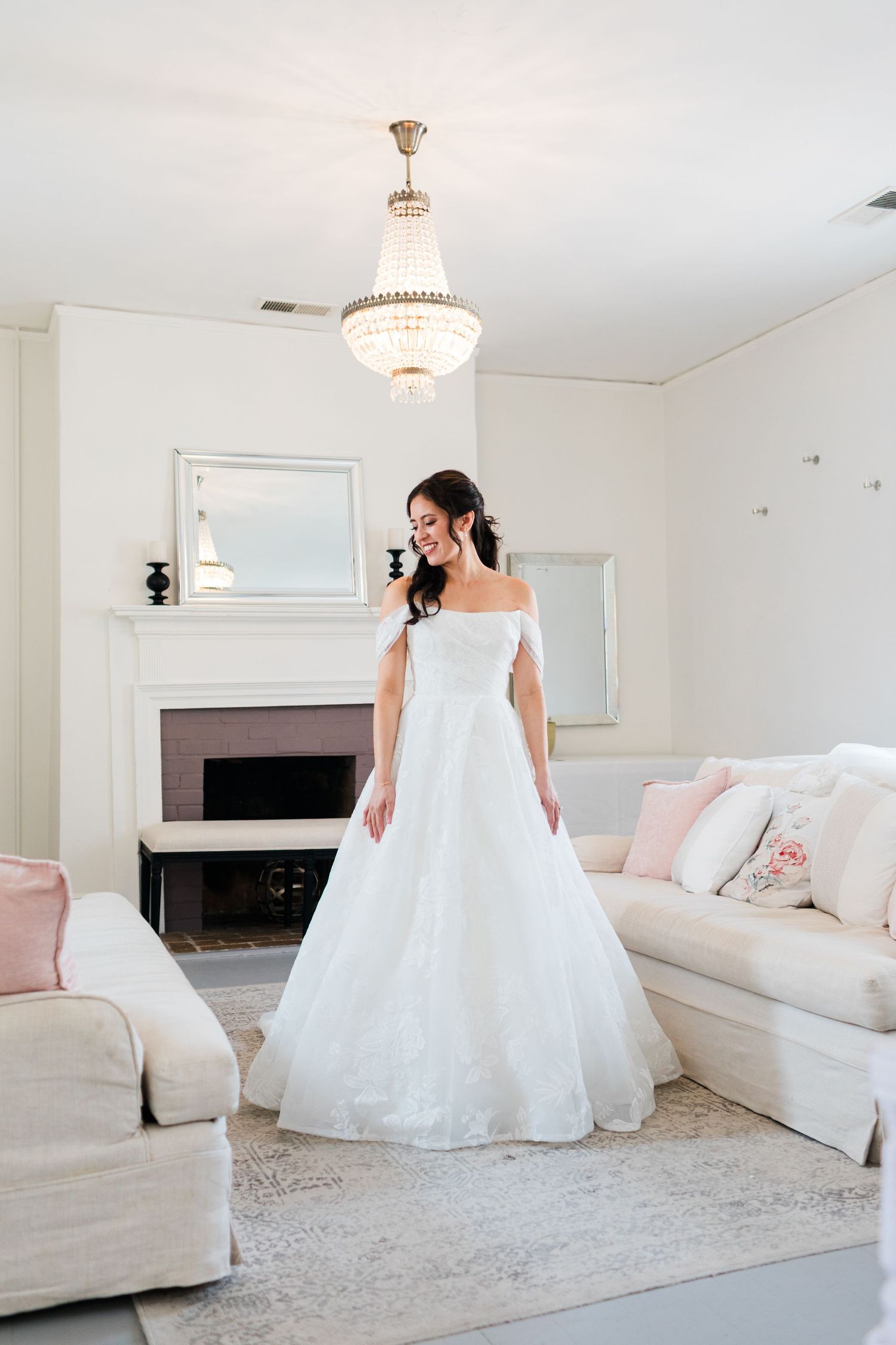 Smiling bride in an off-shoulder white ball gown stands in an elegant white room with chandelier and fireplace.
