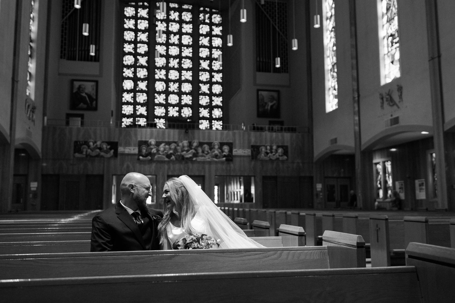 Black and white photo of bride and groom sharing an intimate moment in a church pew with stained glass windows.