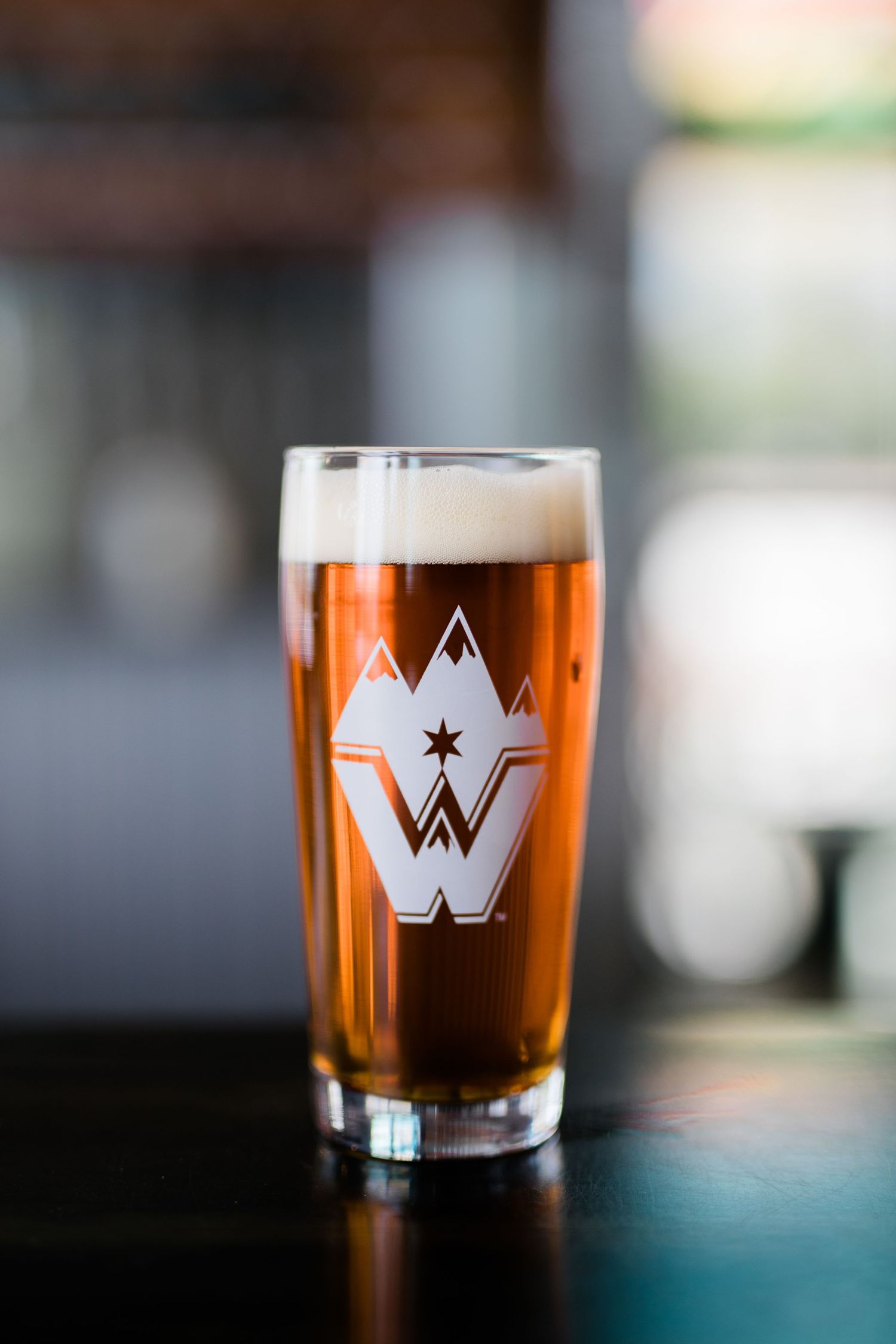 Five identical beer glasses with amber colored beer and white W logo branding sit on a dark surface in diminishing focus.