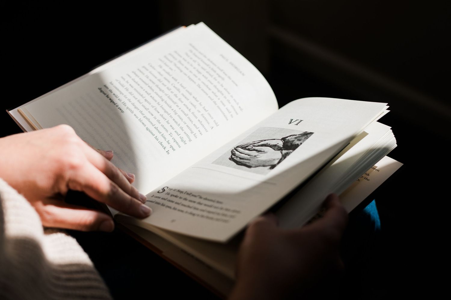 Hands holding open a hardcover book against a dark background, showing pages of text.