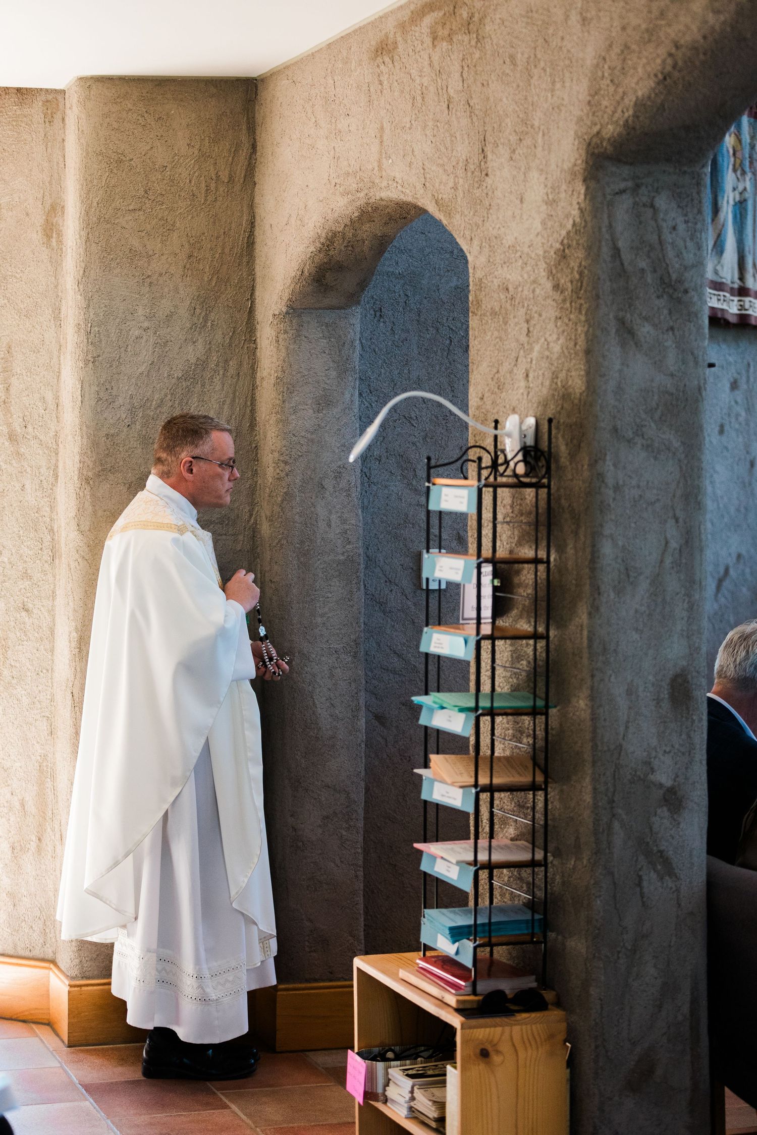 Catholic priest in white vestments stands beside a metal rack of religious pamphlets inside a stone church.