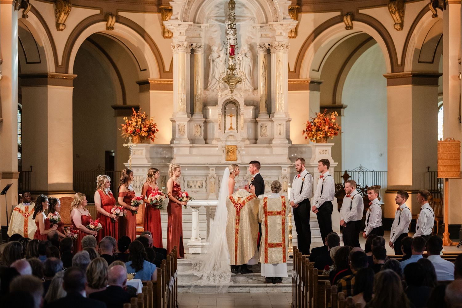 Wedding ceremony at ornate church altar with bride, groom, officiant, bridesmaids in red, and groomsmen.