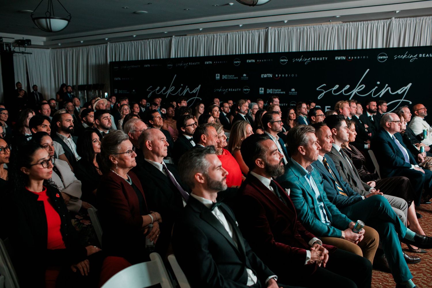 A crowded auditorium filled with formally dressed audience members watching an event in a dark theater setting.