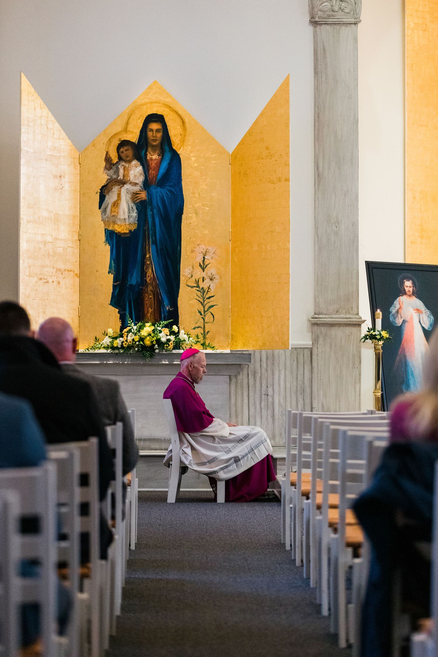 Religious figures kneel in prayer inside a church with yellow walls and rows of white chairs.