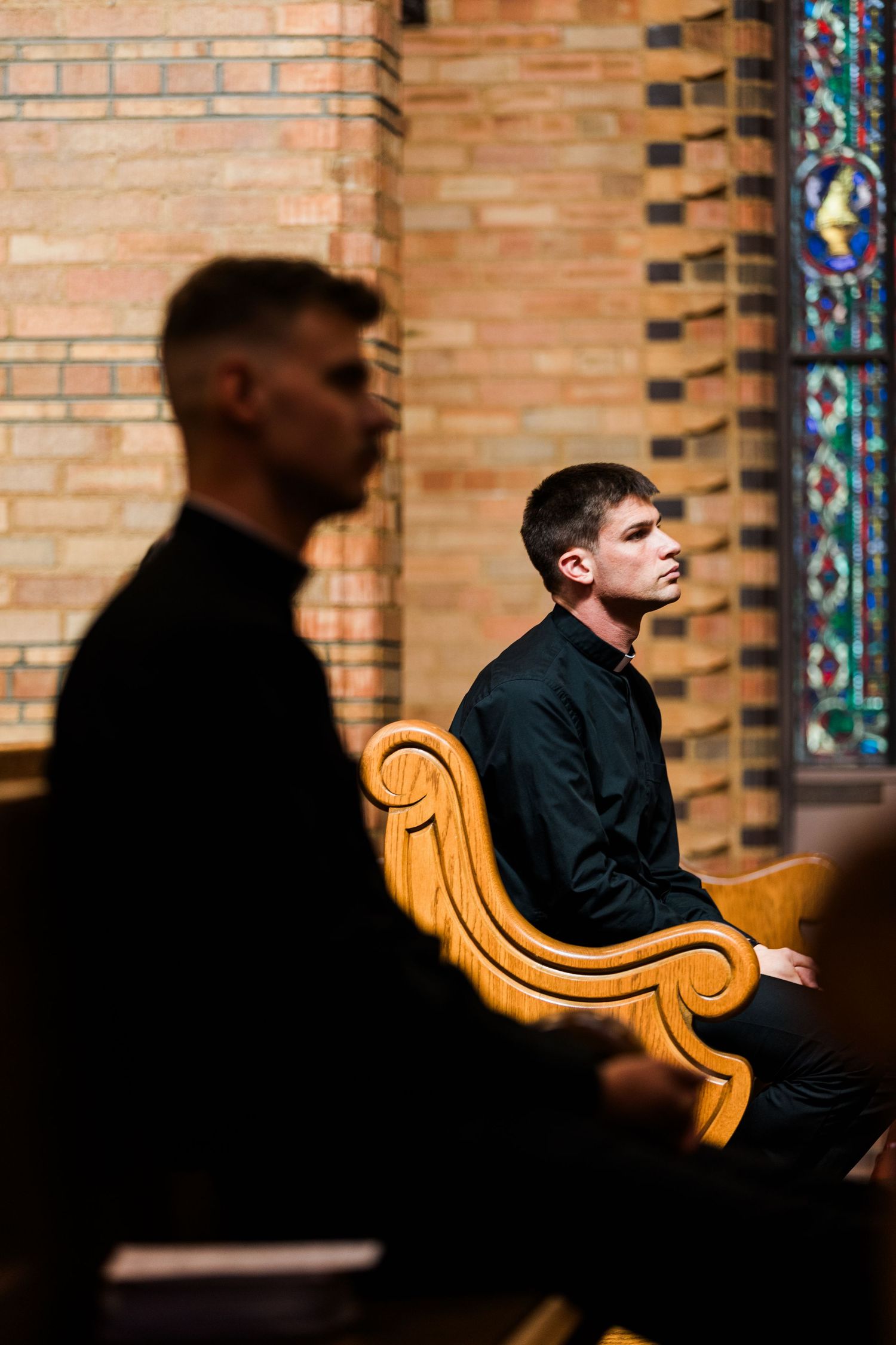 People seated in church pews with ornate stained glass windows and brick walls visible in the background.