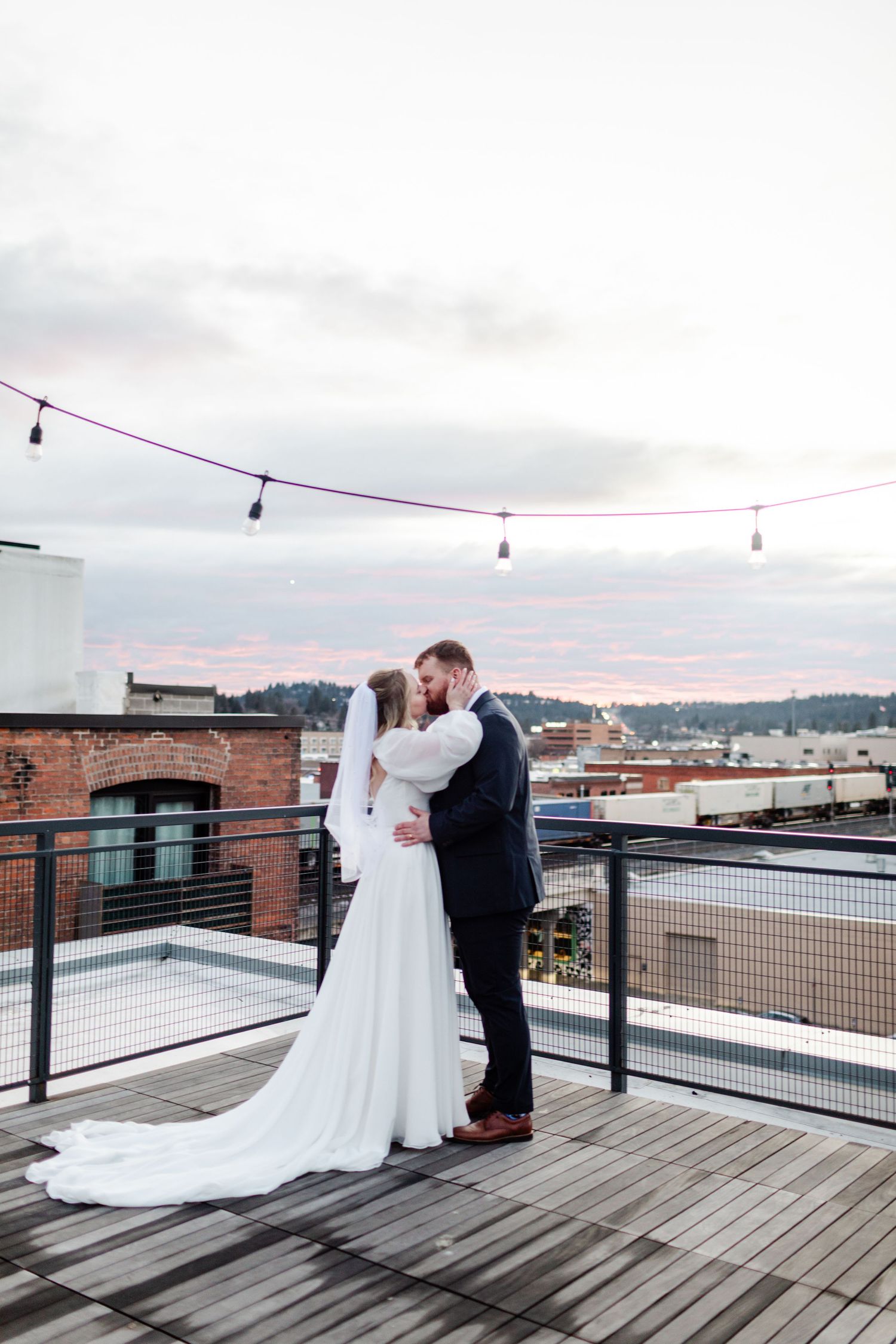 A romantic moment captured on a rooftop deck with string lights and city views in the background.