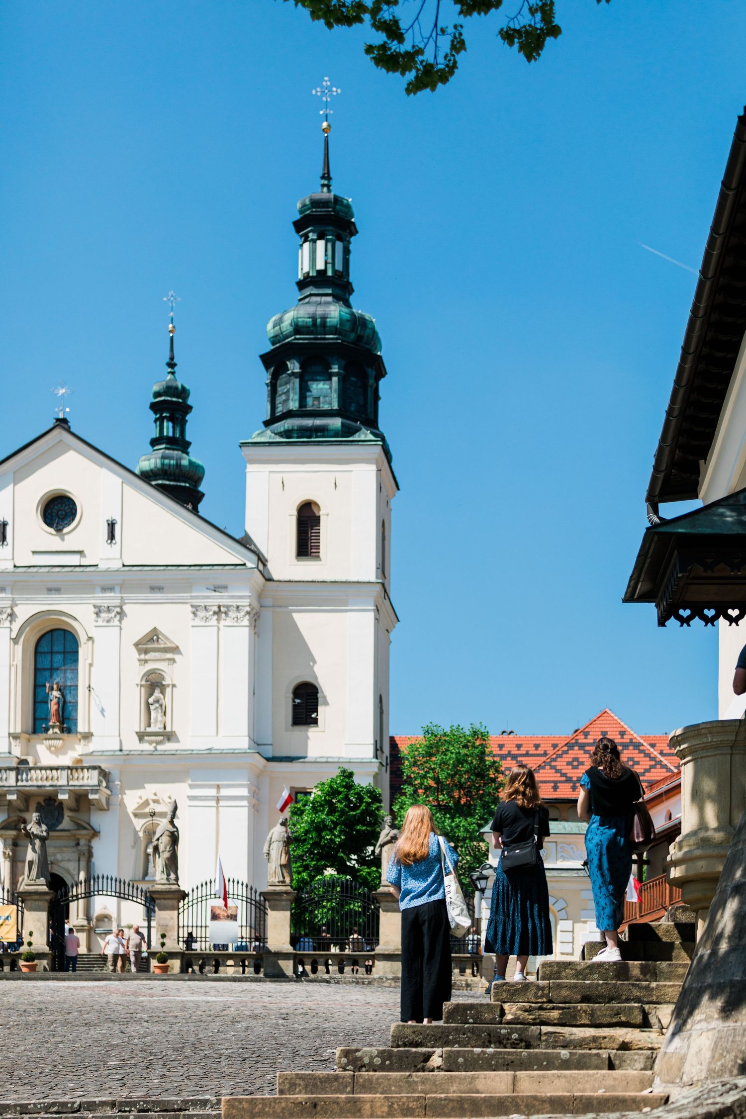 Tourists visiting a historic white Baroque church with green spires under a clear blue sky.
