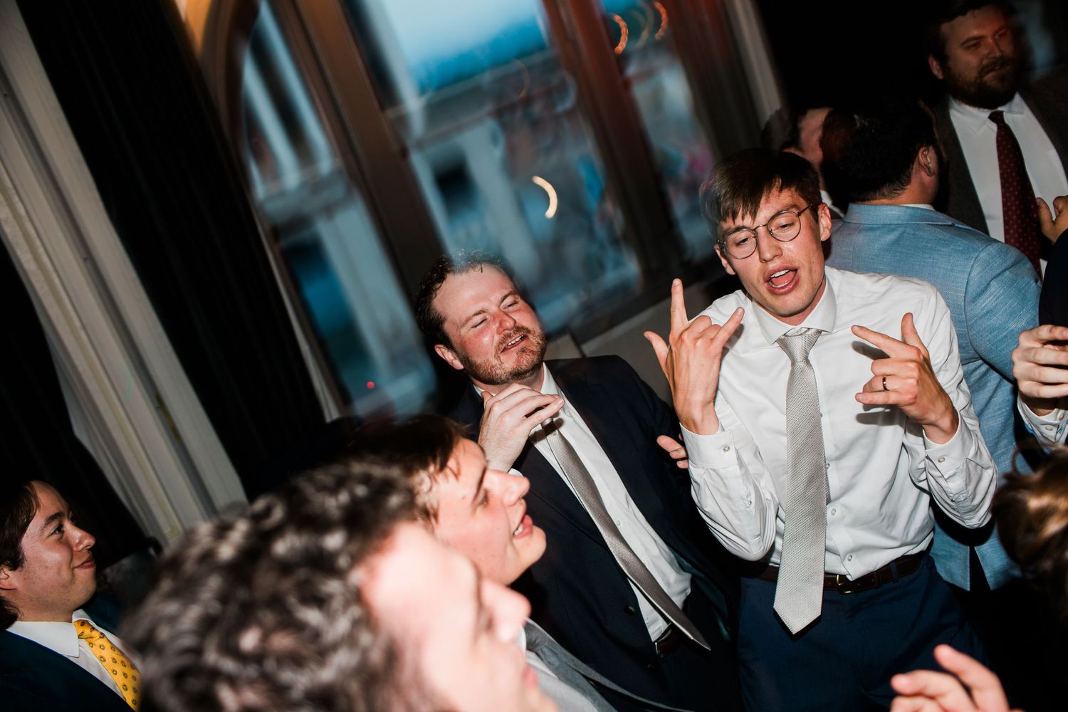 Men in suits celebrating at a wedding reception party, making fun gestures and smiling on the dance floor.