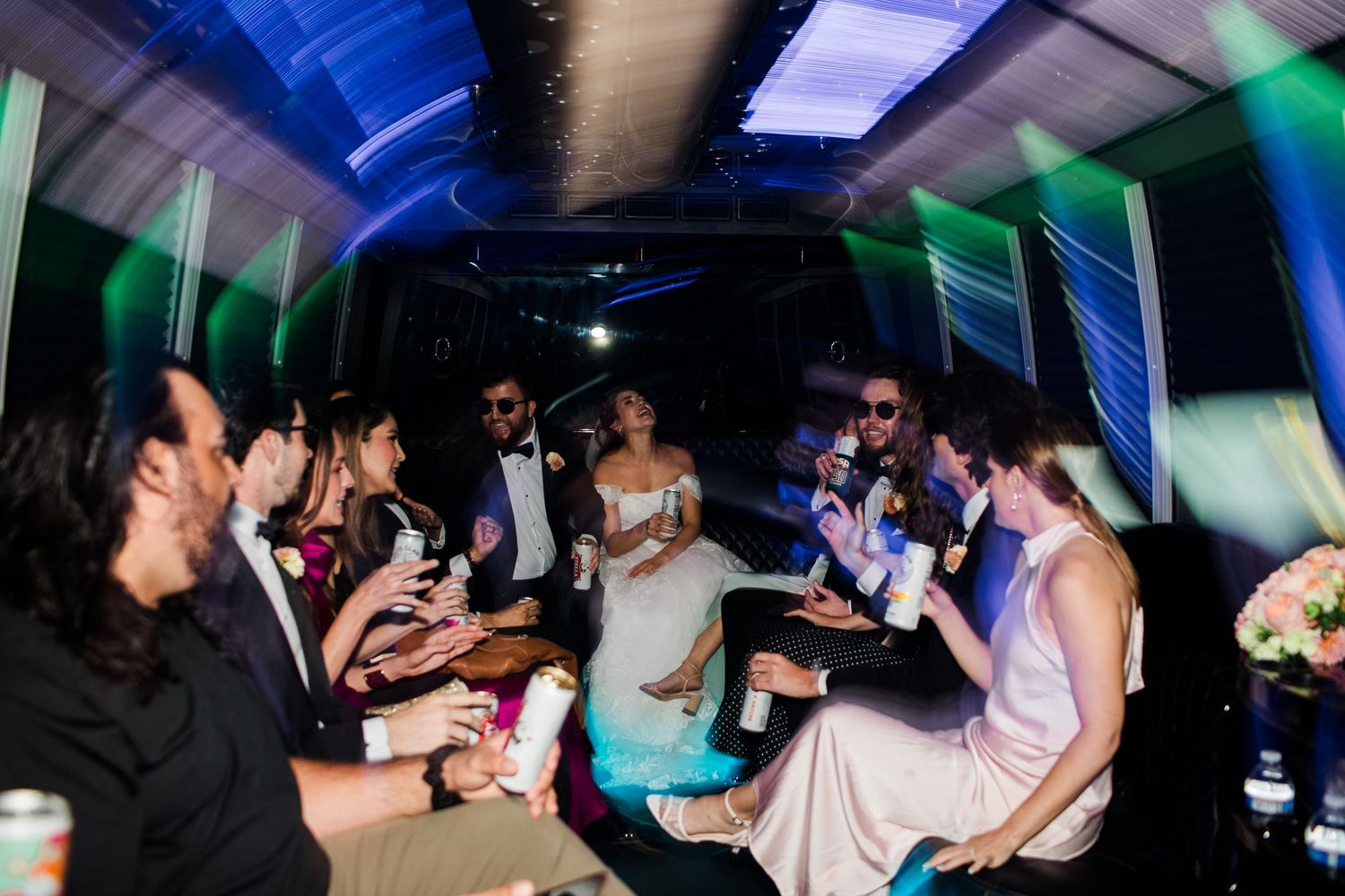 Bride and groom celebrate with wedding party guests inside a decorated party bus with colorful disco lights.