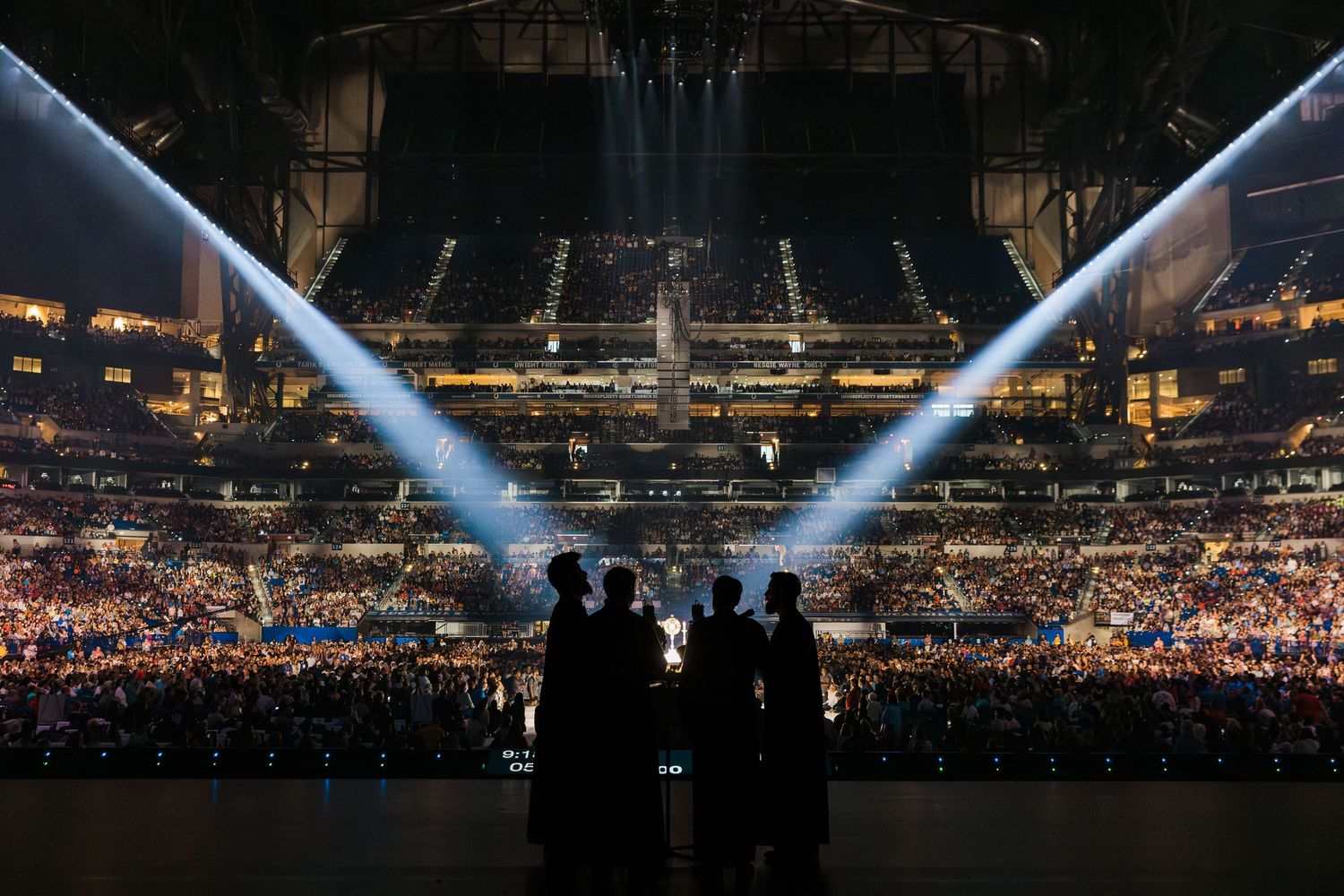Silhouettes of people watching a spectacular arena concert with bright stage lights and packed crowd.