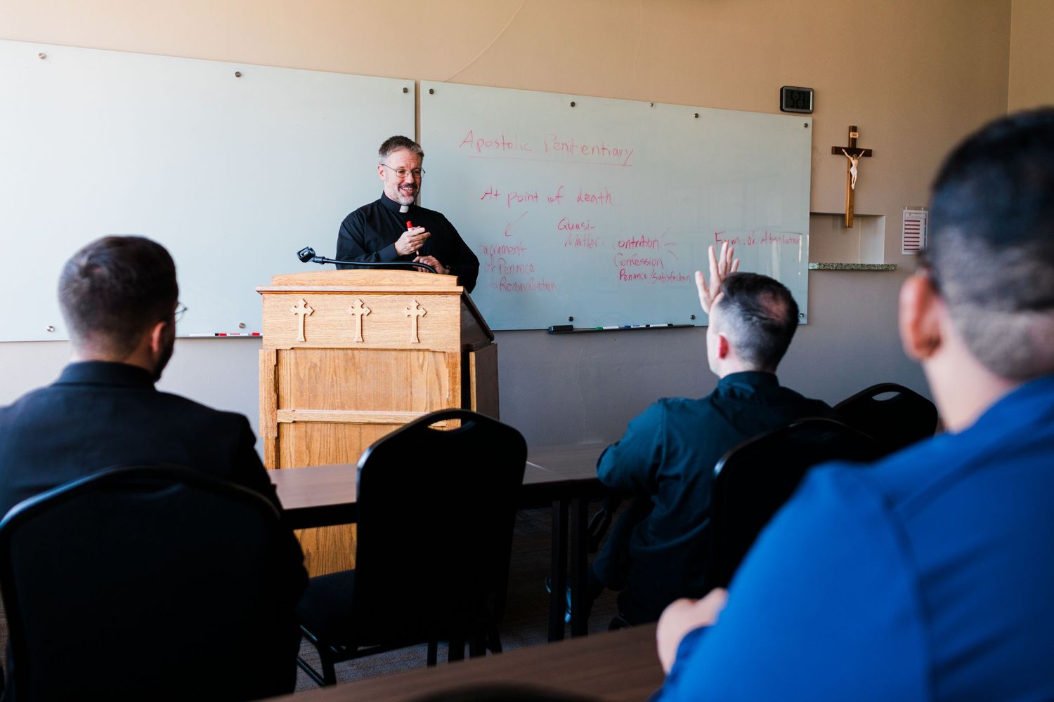 Priest lectures seminary students in a classroom with a whiteboard and crucifix on the wall.