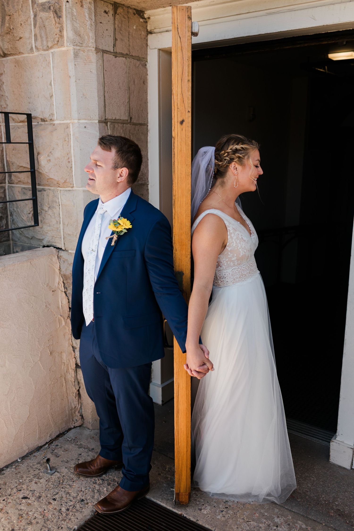 Bride and groom stand back-to-back at a doorway, he in a navy suit with yellow boutonniere, she in a white gown with veil.