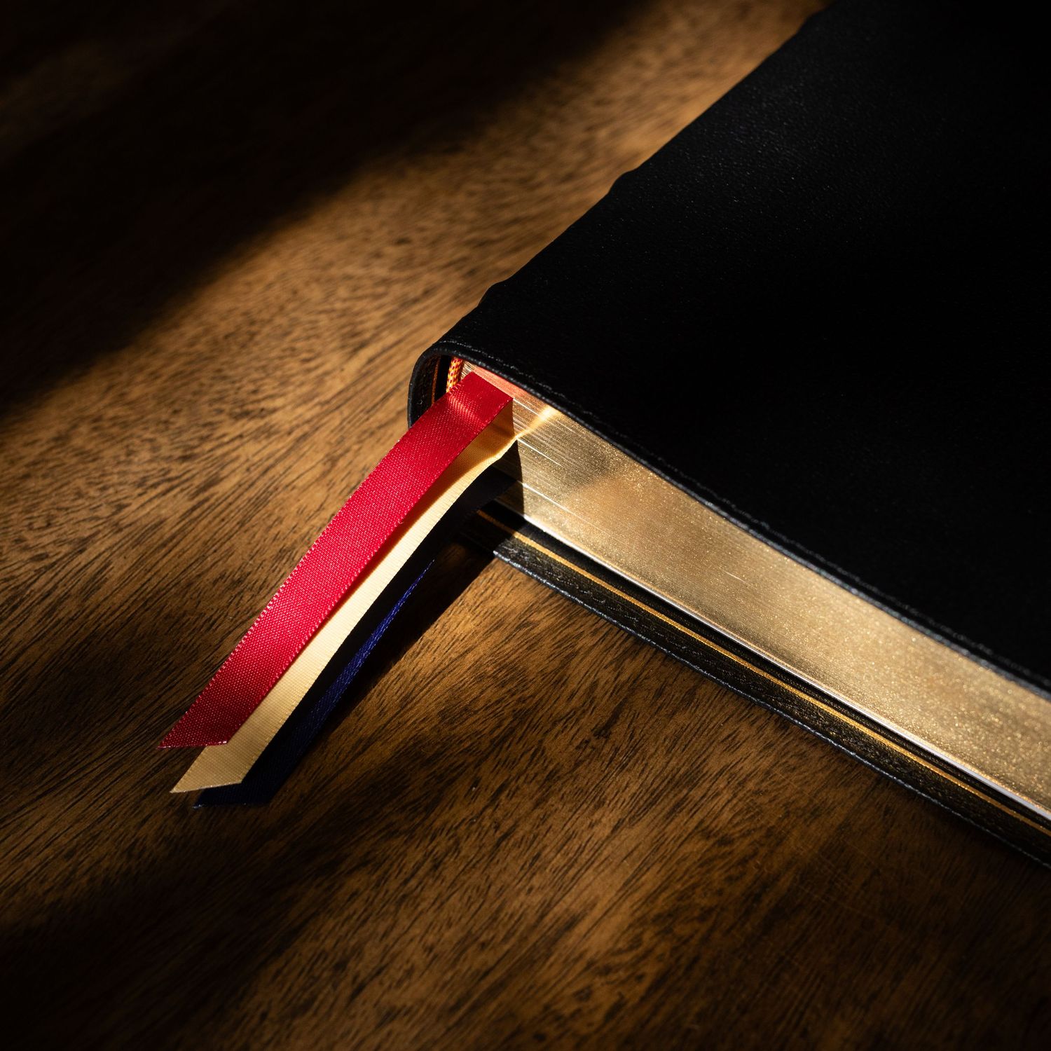 A red bookmark ribbon peeks out from the edge of a dark hardcover book on a wooden surface in dramatic lighting.