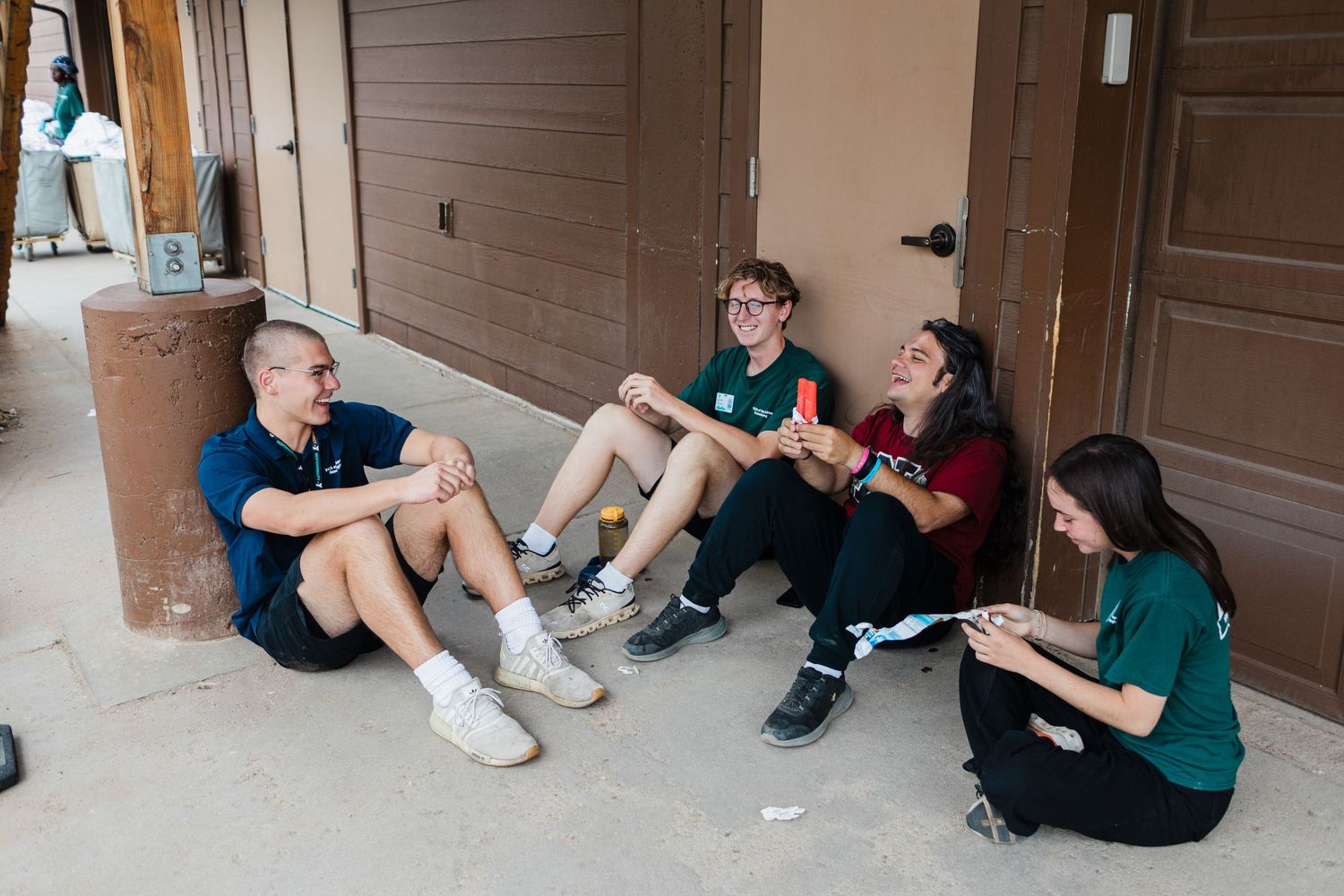 Four teens sit on the ground against a building wall, chatting and relaxing outdoors.